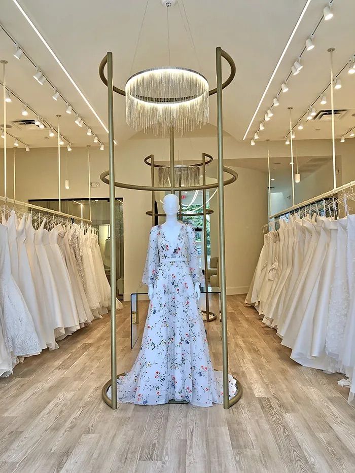 Bridal boutique interior with wedding dresses on racks along both walls, a mannequin wearing a floral long-sleeve wedding gown under a circular chandelier, light wood floor.