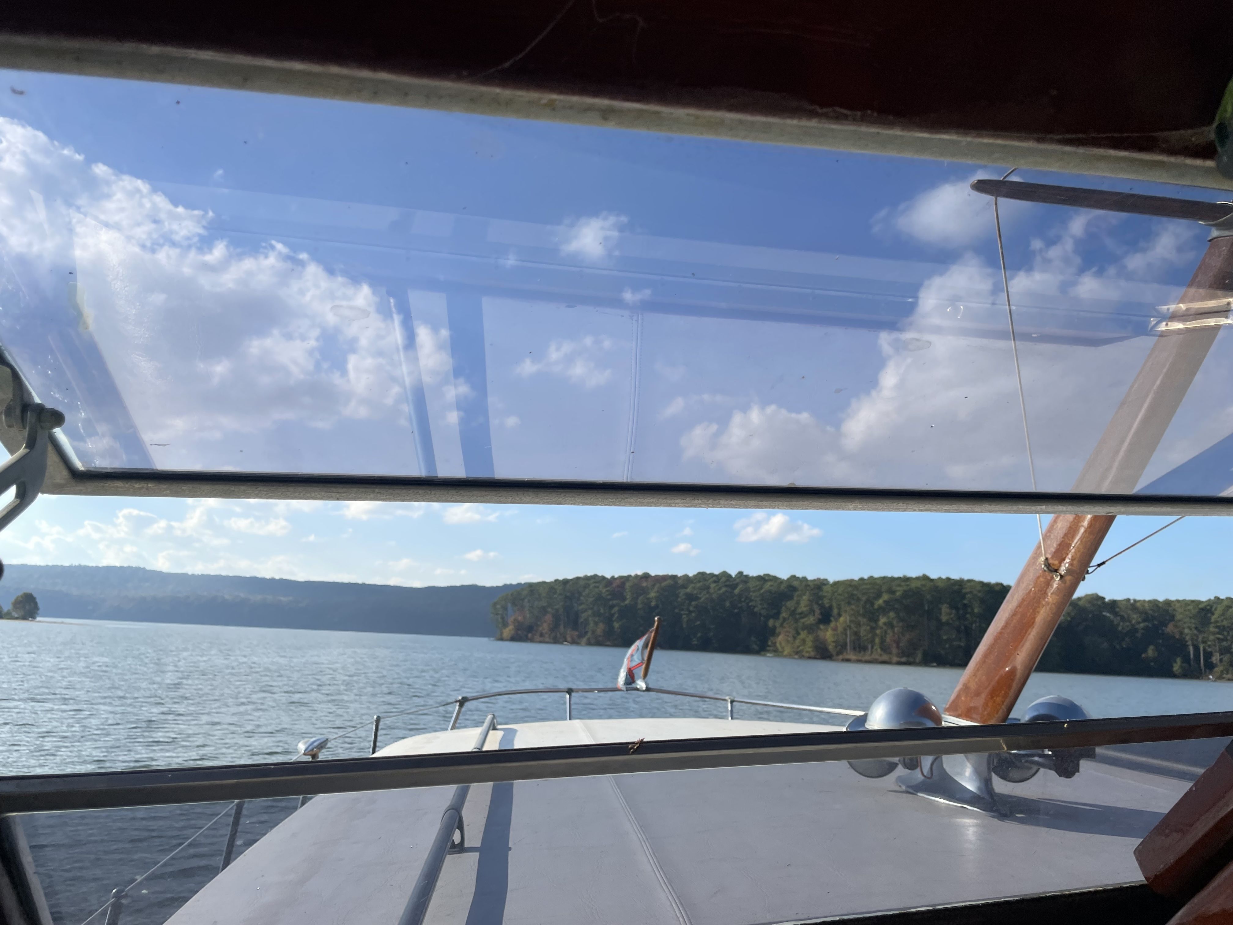 View from a boat cabin: clear blue sky with white clouds over calm water and a tree-lined shore; wooden mast and boat deck visible through the cabin window.