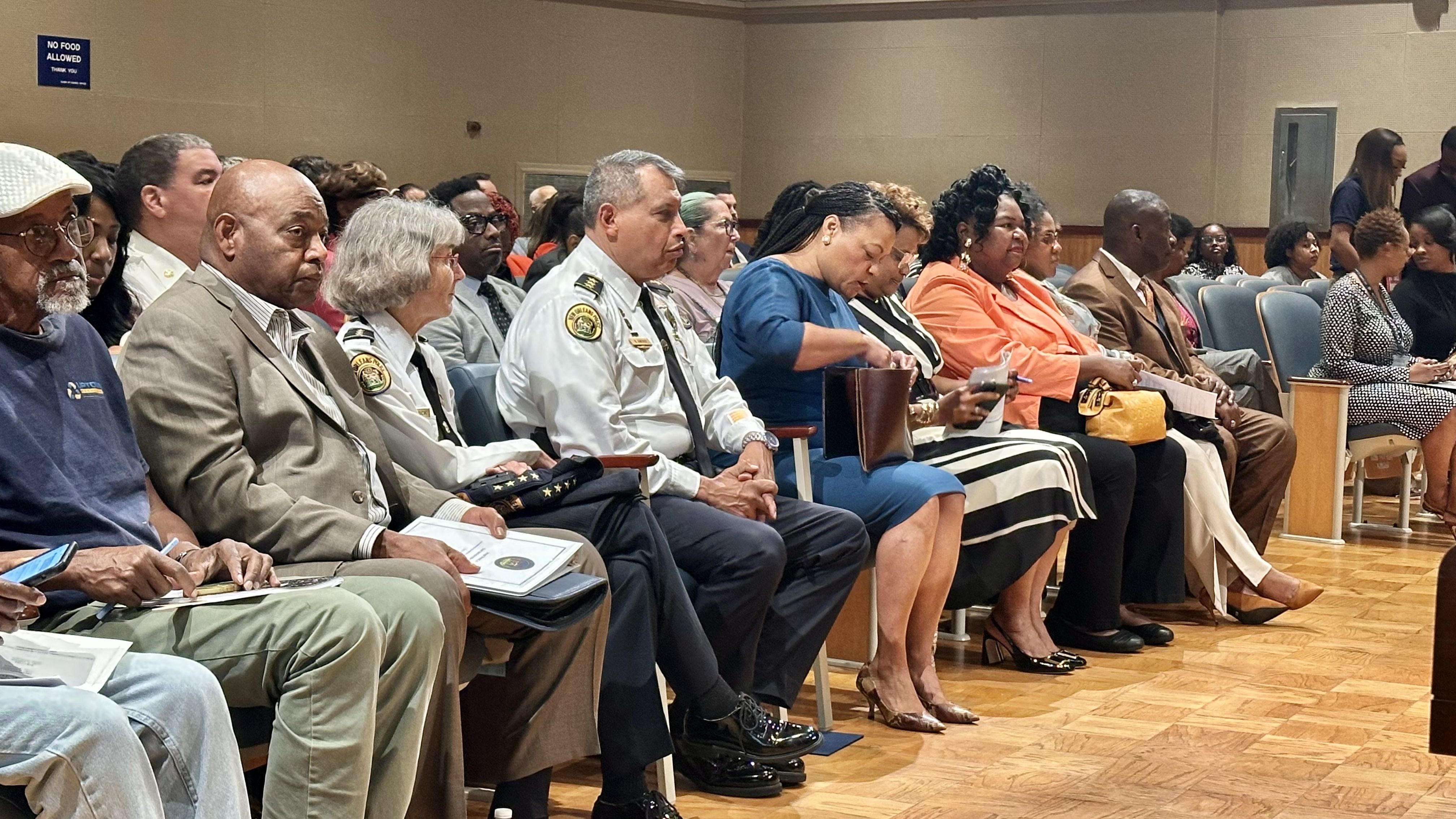 People of diverse ages and backgrounds seated in a row inside a hall. Two officers in white uniforms with badges, others dressed in business or casual attire. A sign reads "No Food Allowed."