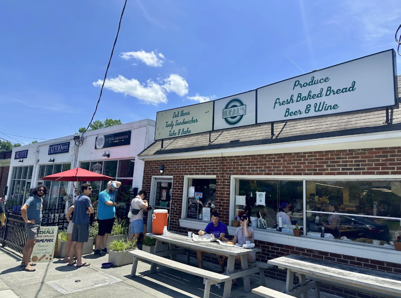 People standing in line outside a brick sandwich shop named Ideal's with signs for deli items, sandwiches, fresh baked bread, beer & wine on a sunny day with a blue sky.