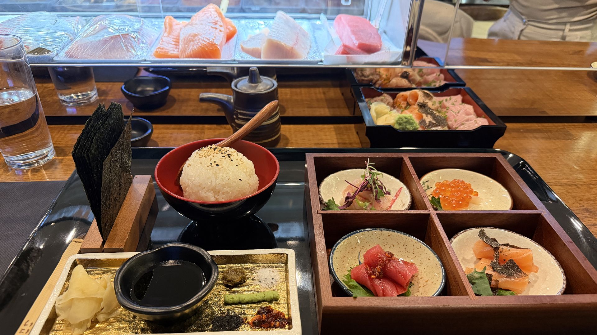 Japanese meal tray with four small dishes of sashimi, a bowl of rice topped with sesame seeds, seaweed sheets, soy sauce, and condiments on a wooden table with a sushi display in the background.