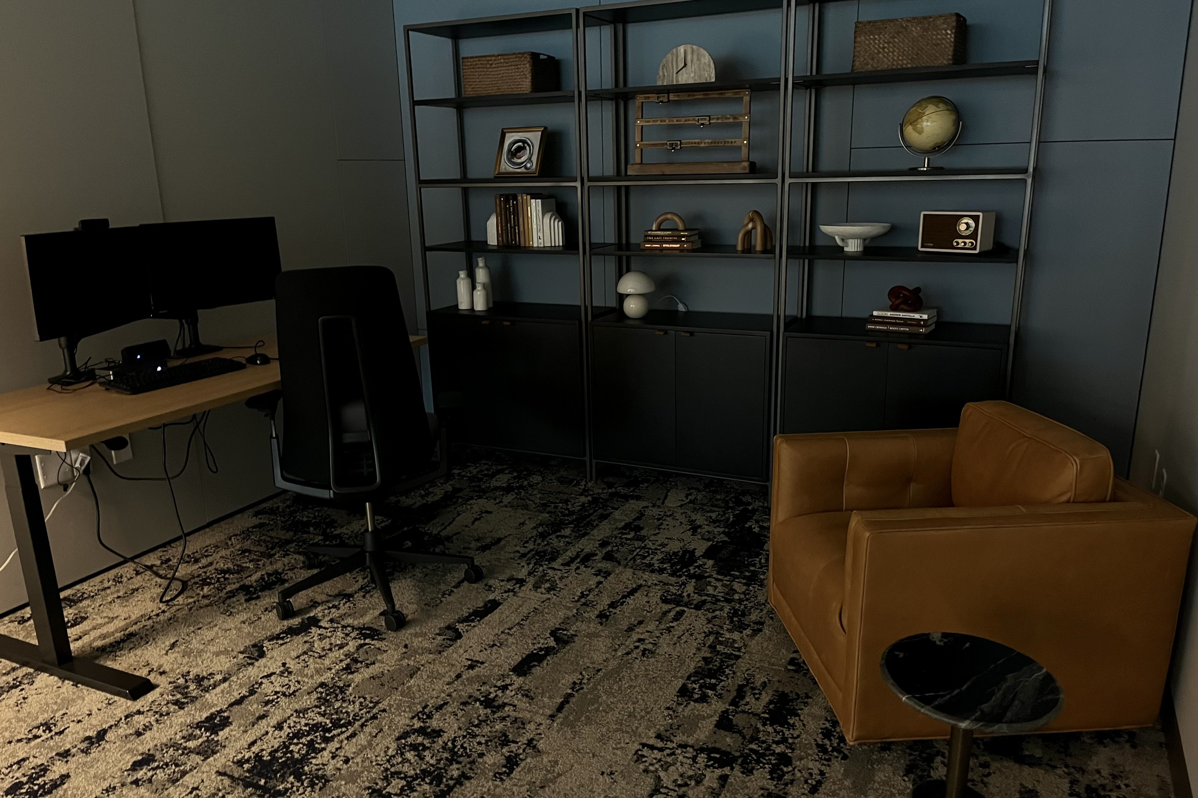 Modern office with black dual monitors on wooden desk, black office chair, brown leather armchair, black shelves with decor items including a globe, books, and vases against blue wall.