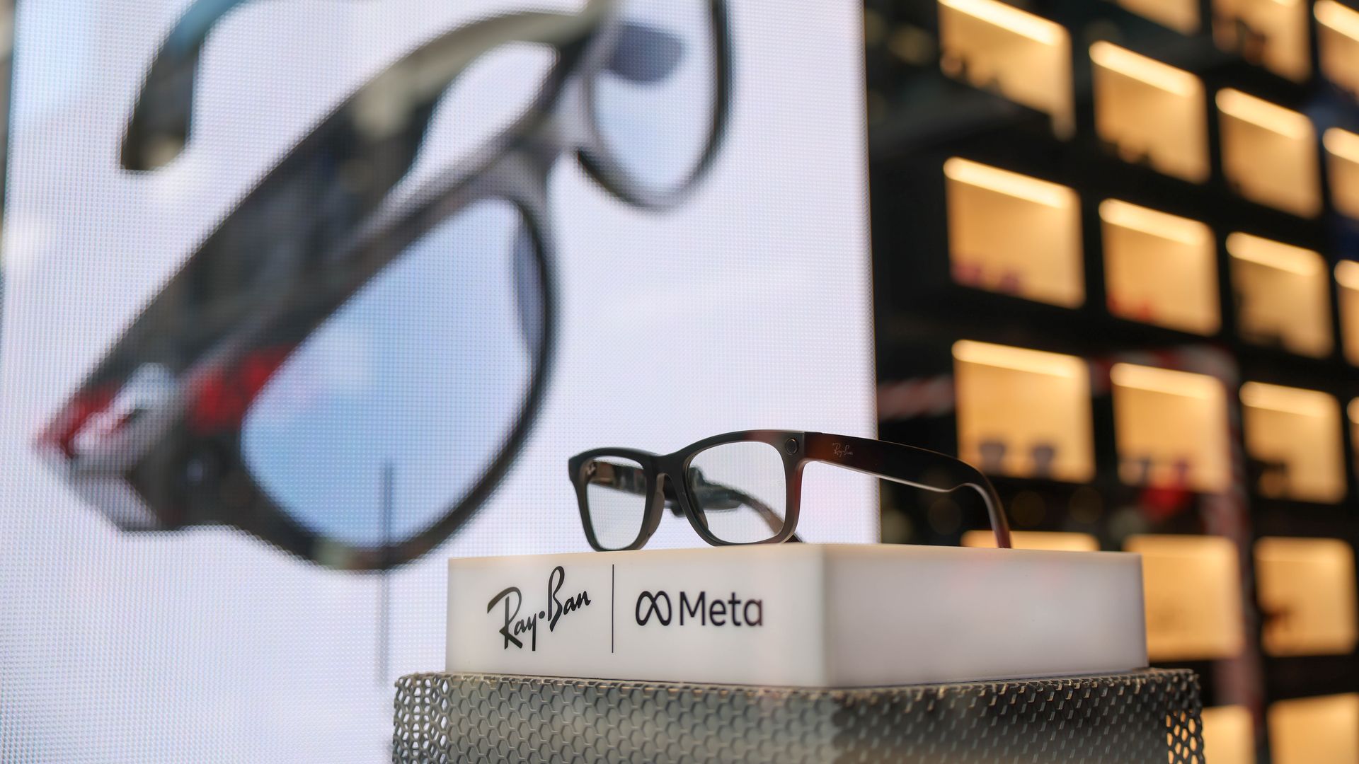 A photo showing a pair of black, squarish glasses displayed on a pedestal that reads: Ray Ban, Meta. Behind it, there is a large image of the glasses on the wall and then lighted up cubbies with glasses in them. 
