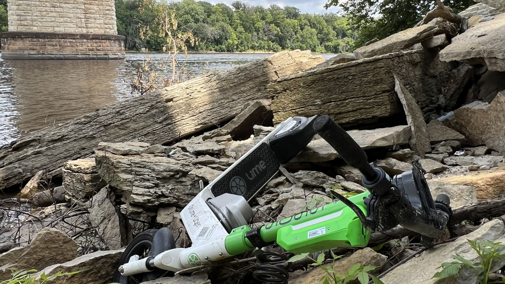 A Lime scooter sitting discarded in a pile of debris underneath a bridge along a river