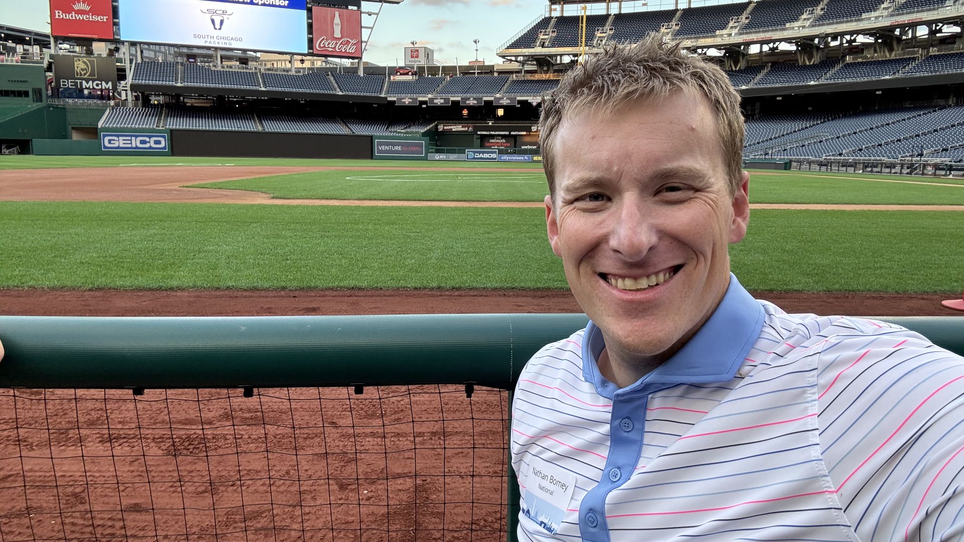 Smiling man taking a selfie at empty Nationals Park baseball stadium, with green field, dirt base paths, and large scoreboard displaying "The Essential Ingredient."