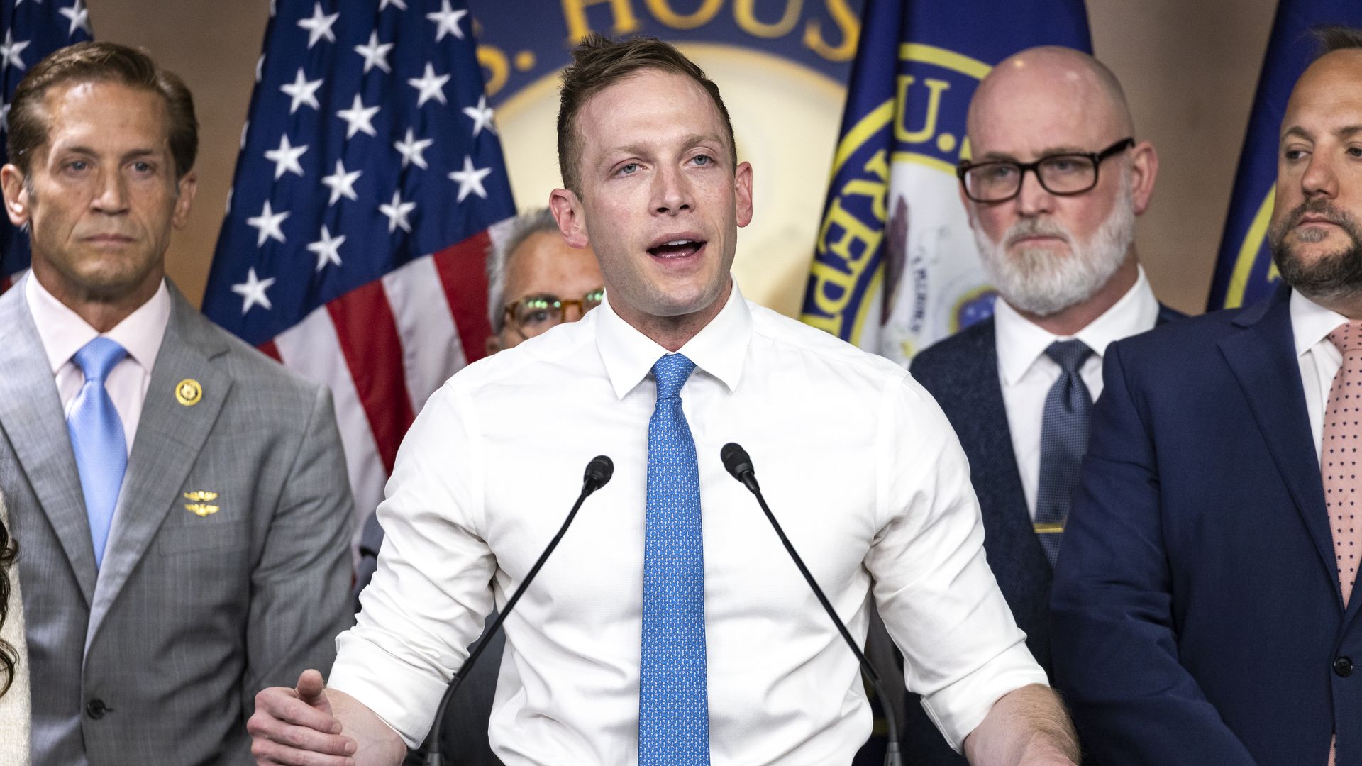 Center: a man in a white shirt and blue tie speaks at a podium with two microphones, flanked by suited men; US flags and a large House seal are in the background.