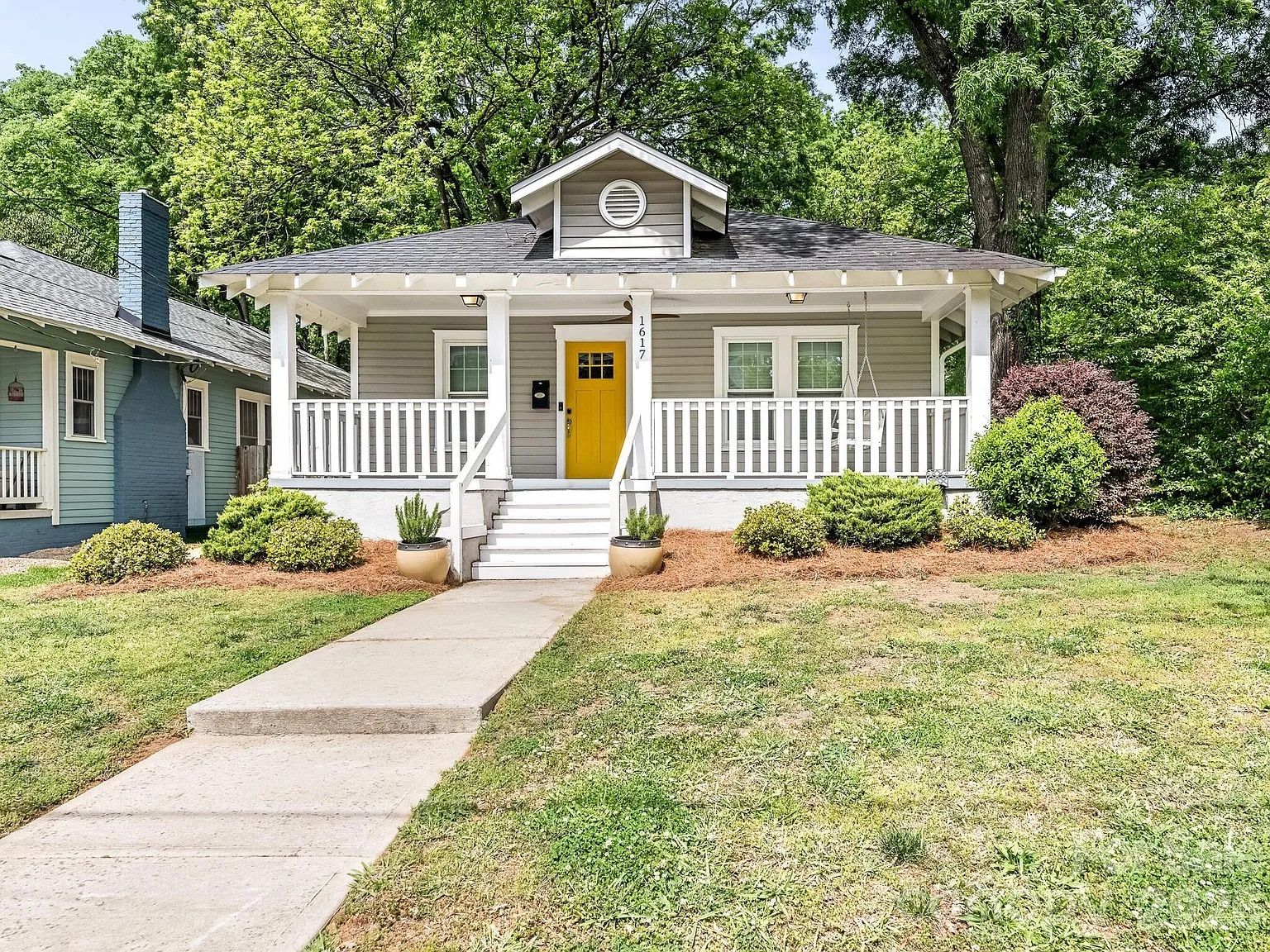 A charming single-story gray cottage with a bright yellow door and white front porch. White railings, steps, potted plants, green lawn, mulch beds, and large trees in the background.