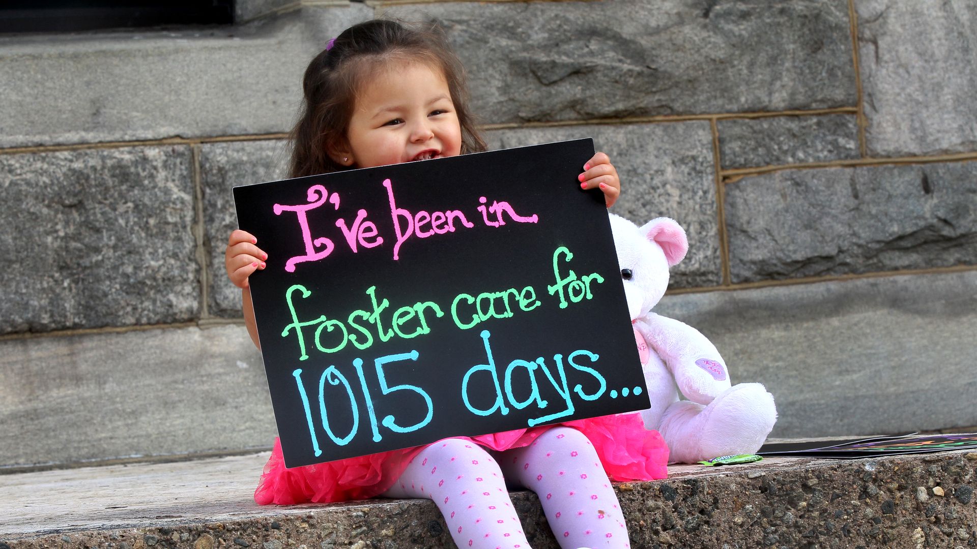 Alexandra Rubio holds a sign that reads, "I've been in foster care for 1,015 days" while sitting on the steps of a Massachusetts courthouse before her adoption proceedings.