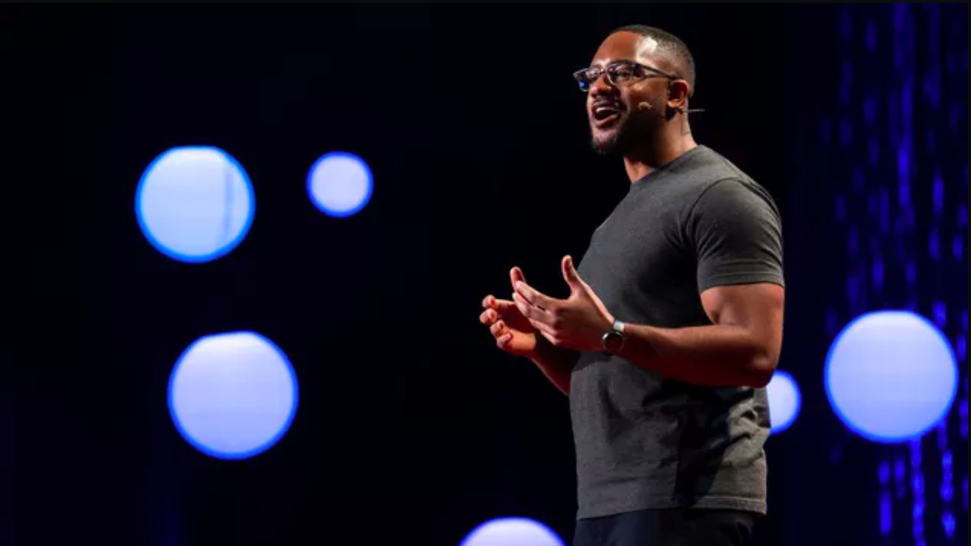 In this image, Brandon Anderson stands on a stage with blue circles in the background. 