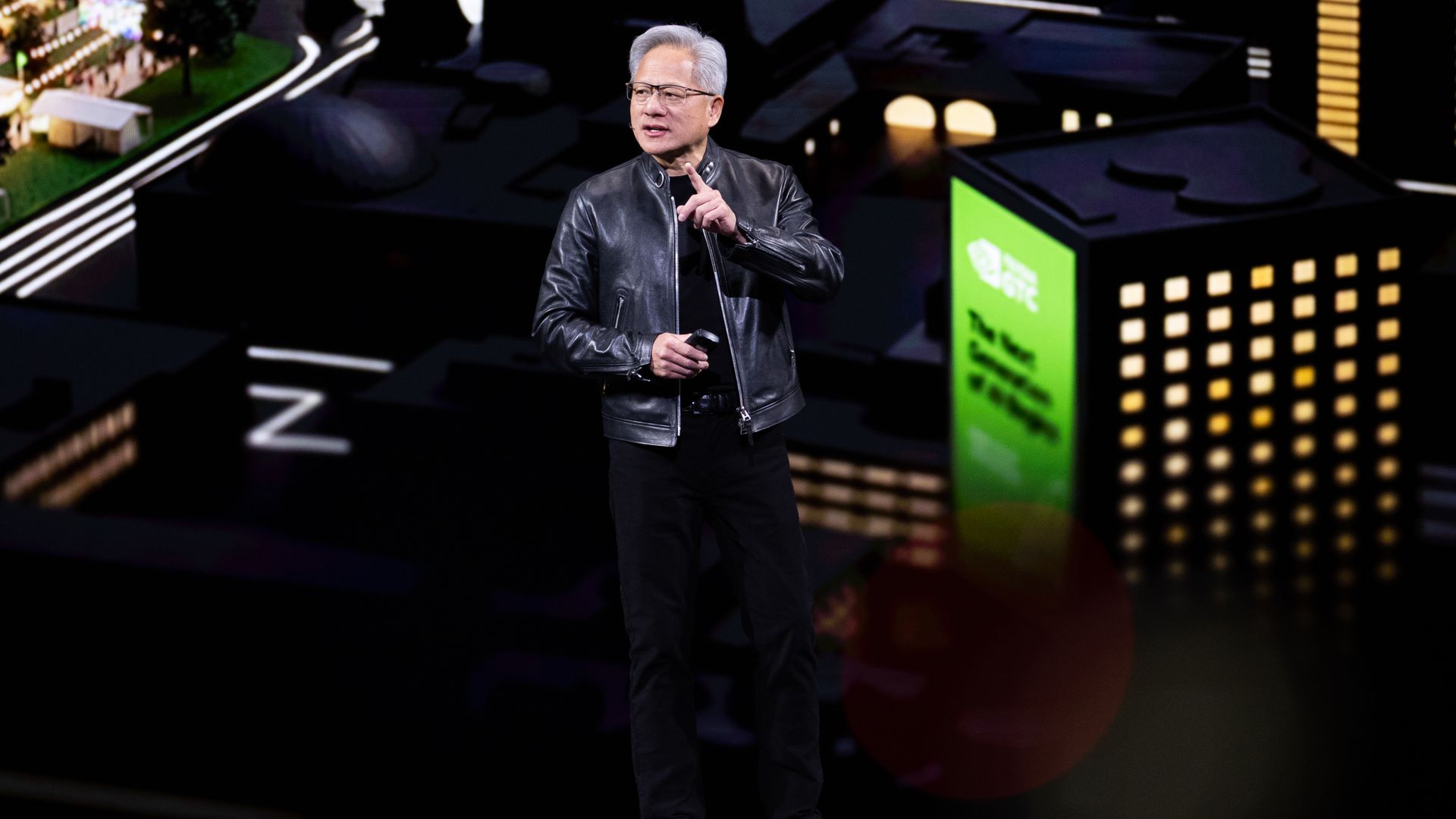 A personin a black leather jacket and black pants speaks into a microphone on a dark stage, with a neon cityscape backdrop of lit buildings behind him.