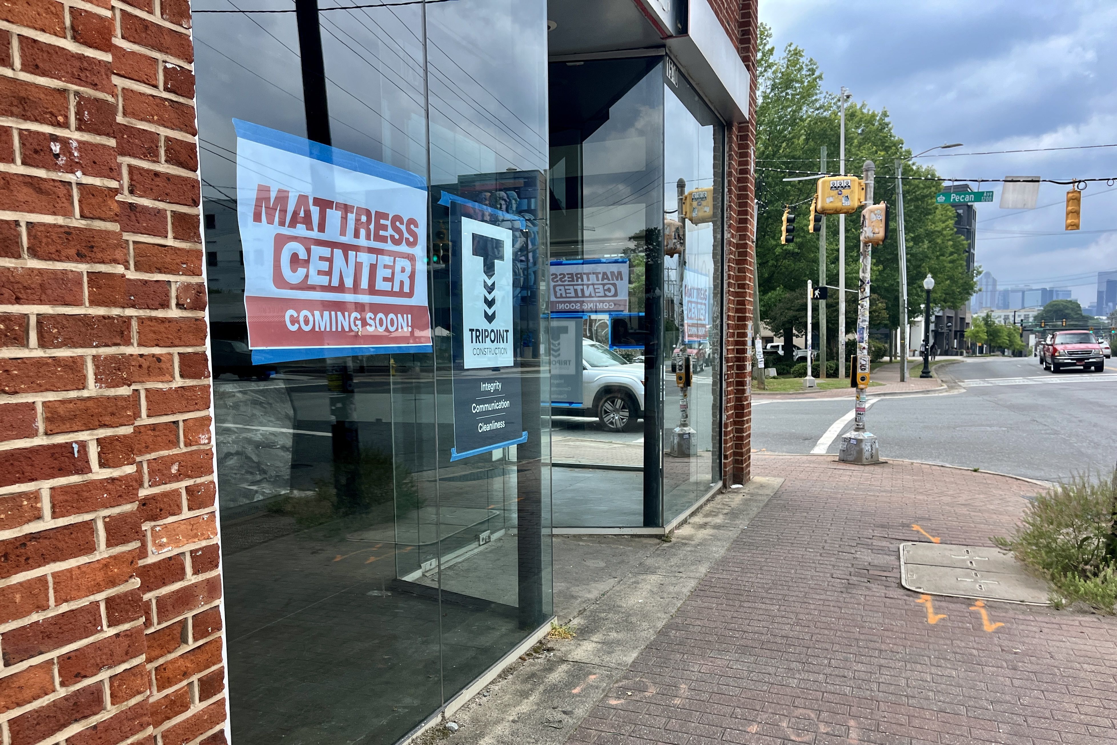 Brick storefront with glass windows displaying a red Mattress Center Coming Soon! sign on blue. Reflections of street, yellow traffic signals, parked cars, trees, and a cloudy sky.