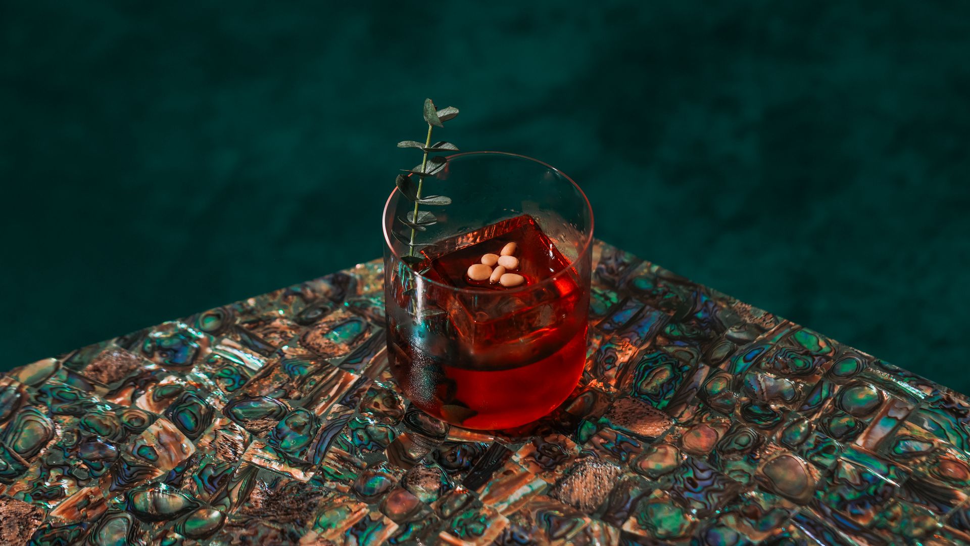 Red cocktail in a clear glass with a large ice cube, garnished with a small green sprig and white seeds, placed on a shiny, multicolored abalone shell patterned table.