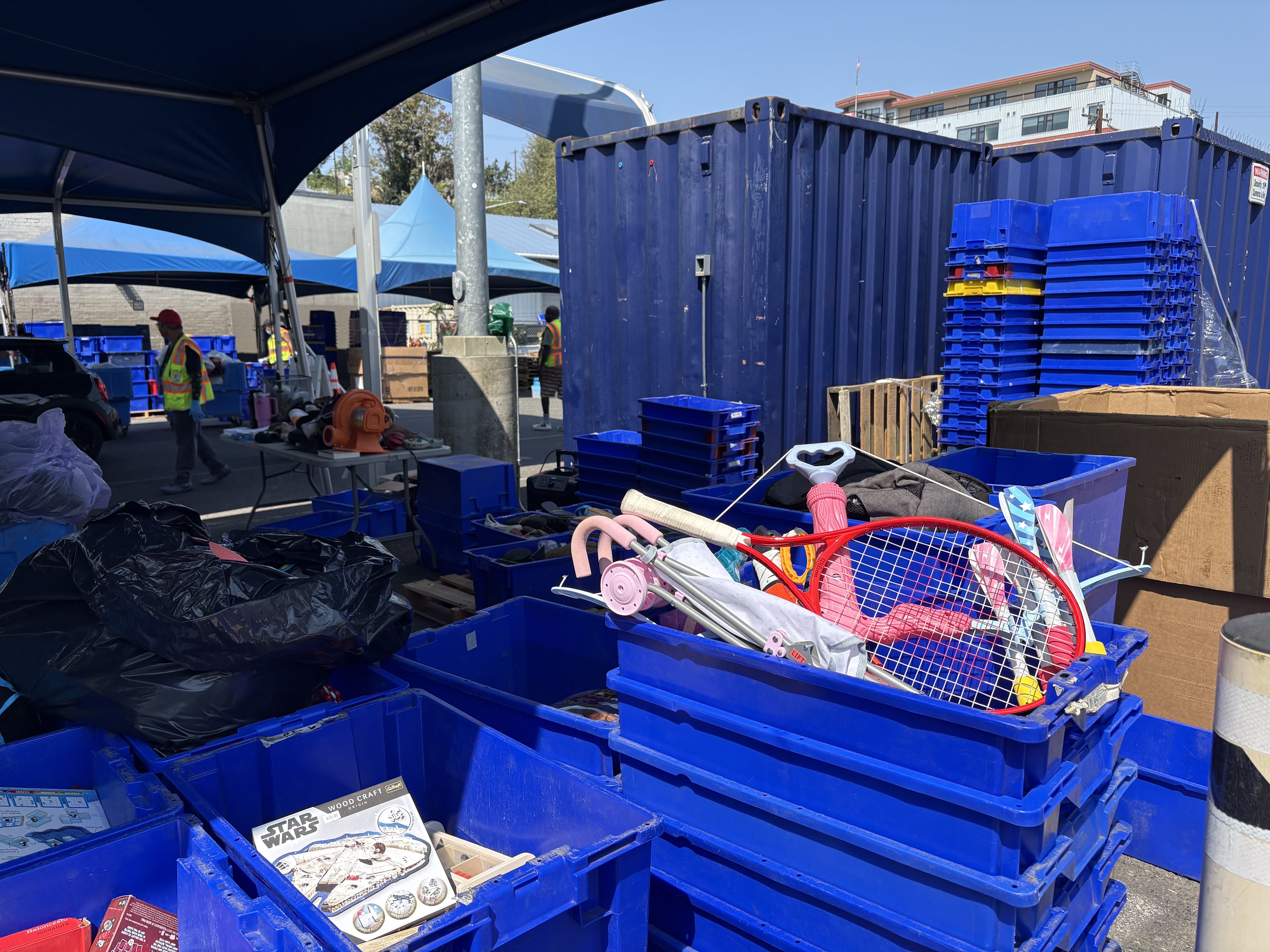 Industrial outdoor yard with blue dumpsters and stacked blue crates under large canopies; workers in high-visibility vests near equipment, pallets, and a car, with a building in the background.