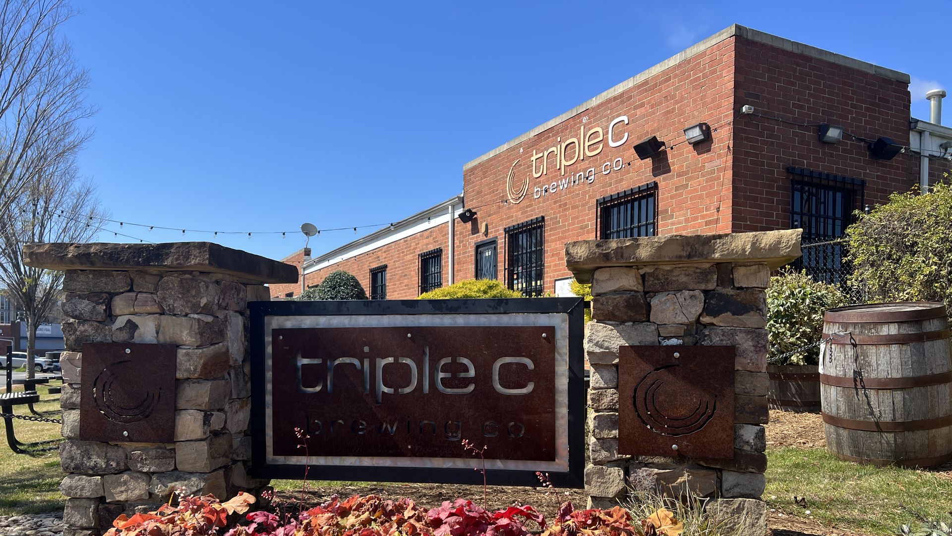 Outdoor view of Triple C Brewing Co. building with brick exterior, metal sign on stone pillars, colorful autumn plants, wooden barrel, and string lights under a clear blue sky.
