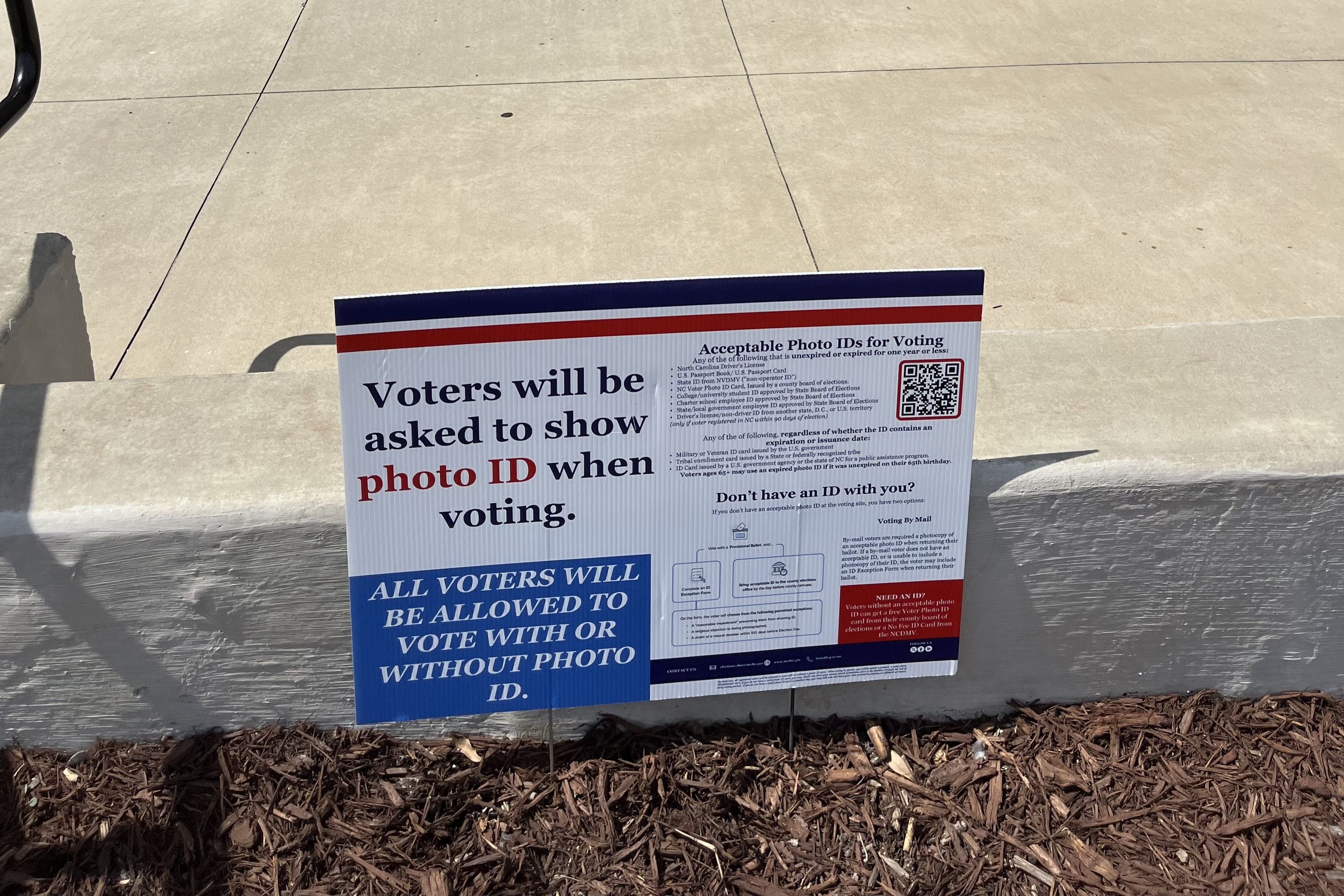 Outdoor sign on a low concrete barrier beside a sidewalk; blue and red stripes at top, bold text about voters showing photo ID when voting, with mulch and wood chips on the ground.