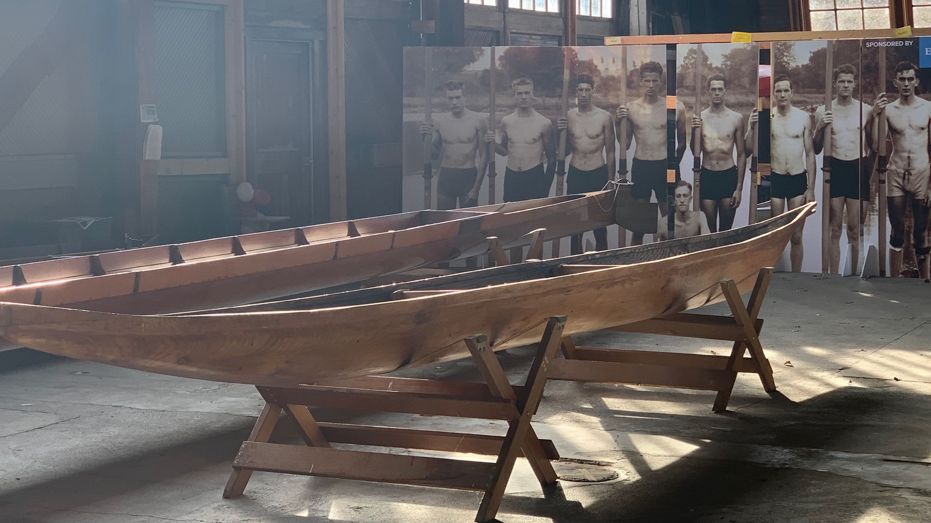 Wooden racing shells in front of pictures of the Olympic Gold Winning team from "The Boys in the Boat."
