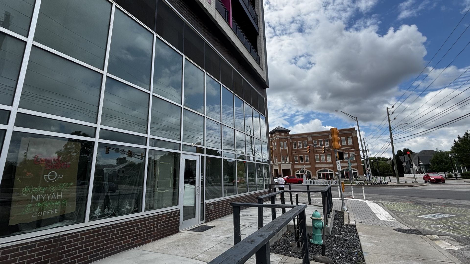 Low-angle street scene showing a large glass-front building with metal grids on the left, brick base and railing. Across the street: a red-brick building, traffic lights, power lines, and a blue sky with clouds.