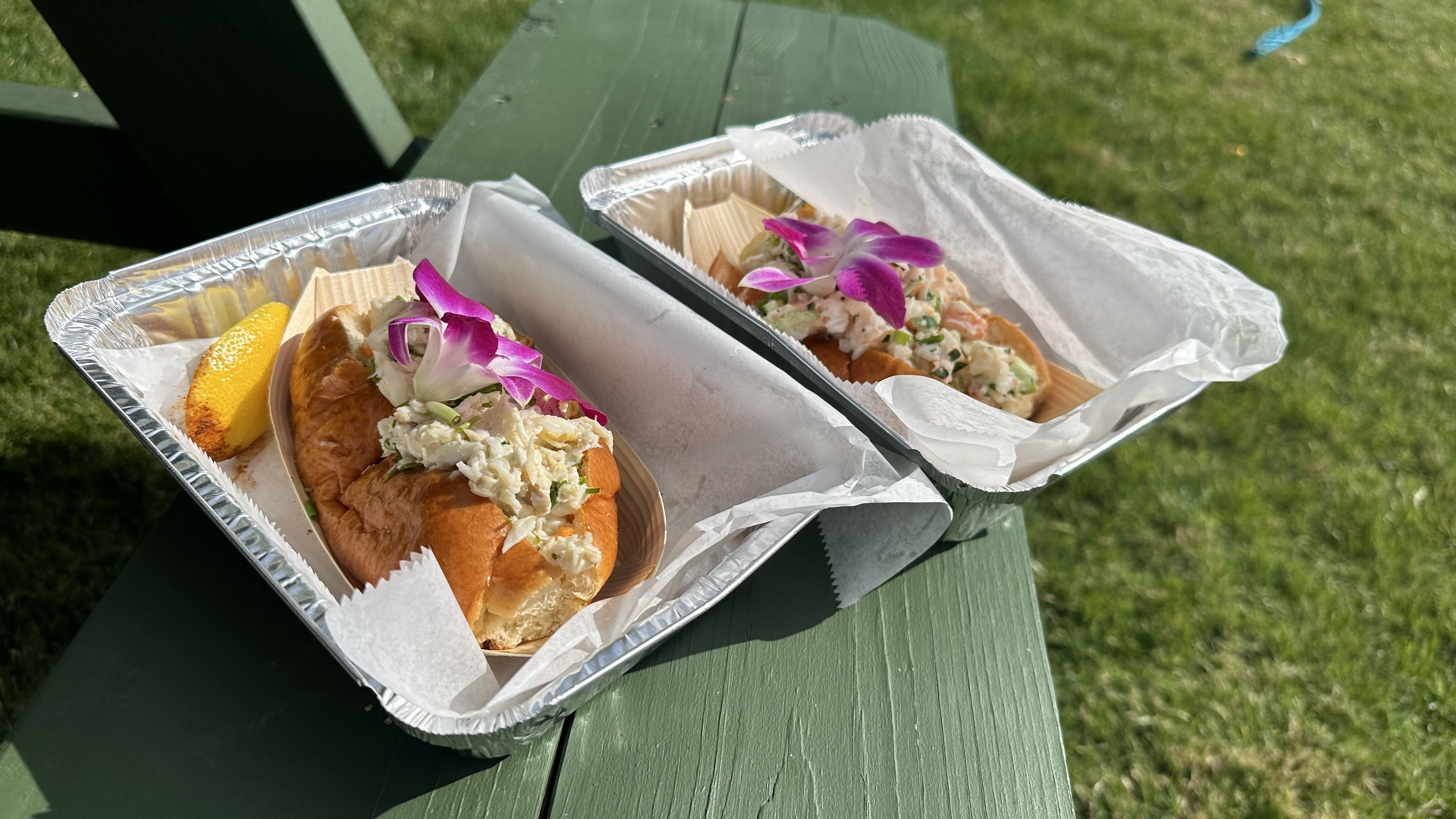 Two seafood salad sandwiches sit in aluminum tins on a park bench.