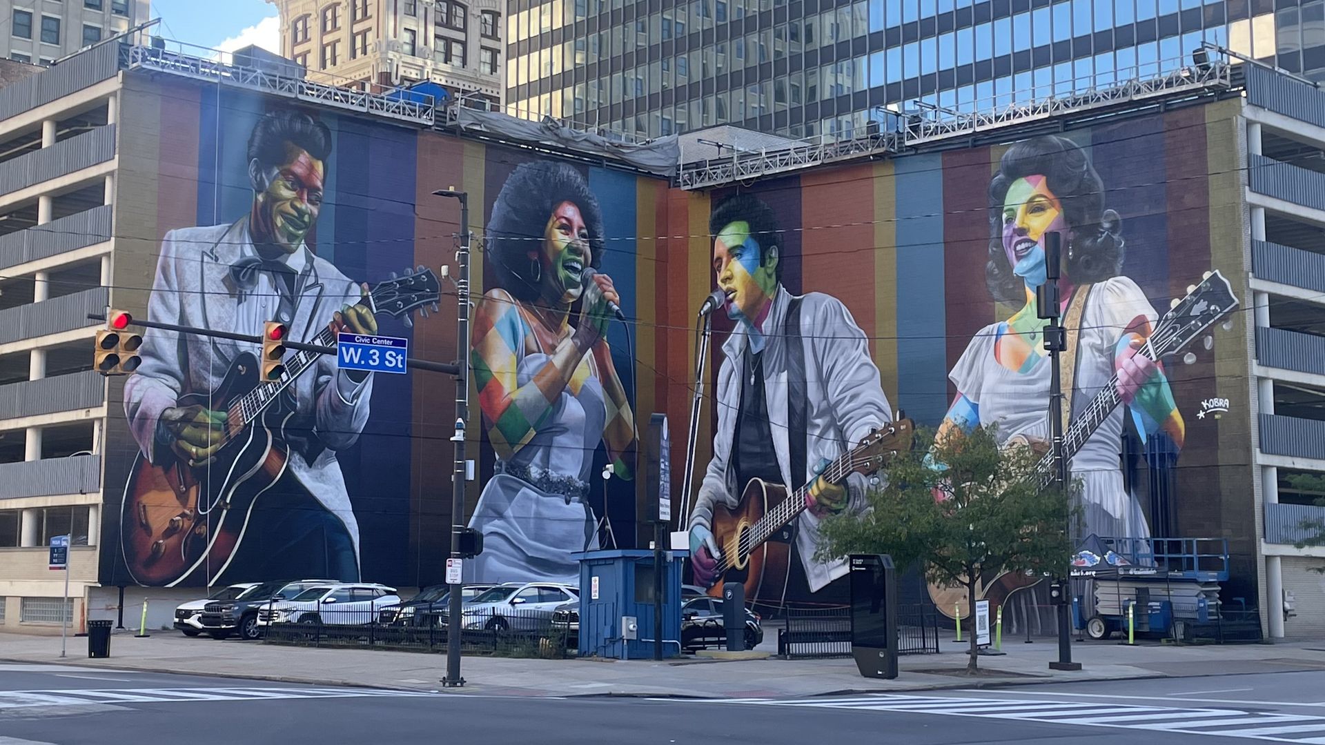 A large mural on a city building depicts four colorful musicians with guitars and a microphone, painted in rainbow hues, set against a backdrop of tall office towers and street signs.