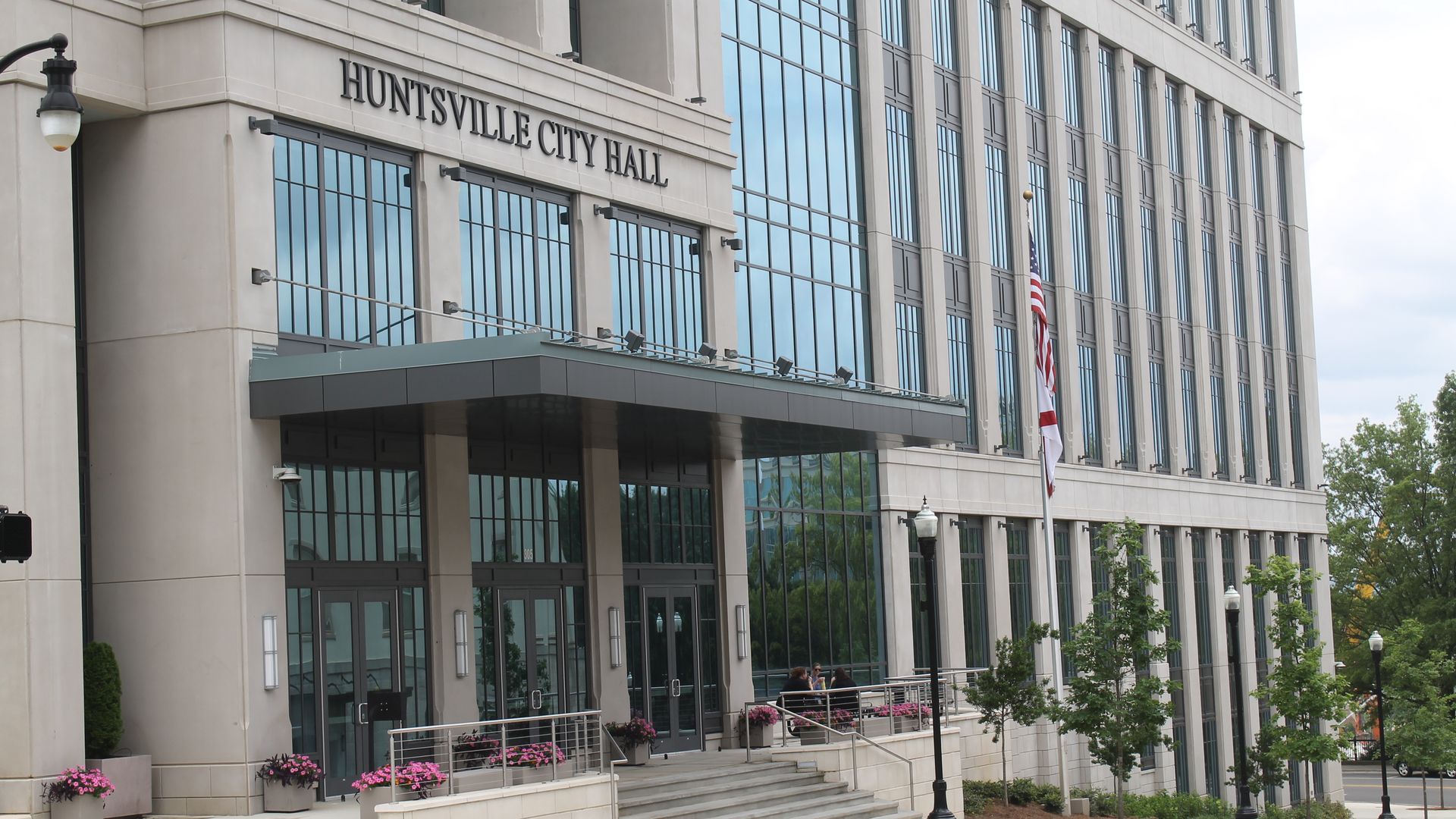 Exterior of Huntsville City Hall, a modern multi-story building with large glass windows, beige facade, pink flowers in pots, and an American flag on a flagpole near the entrance.