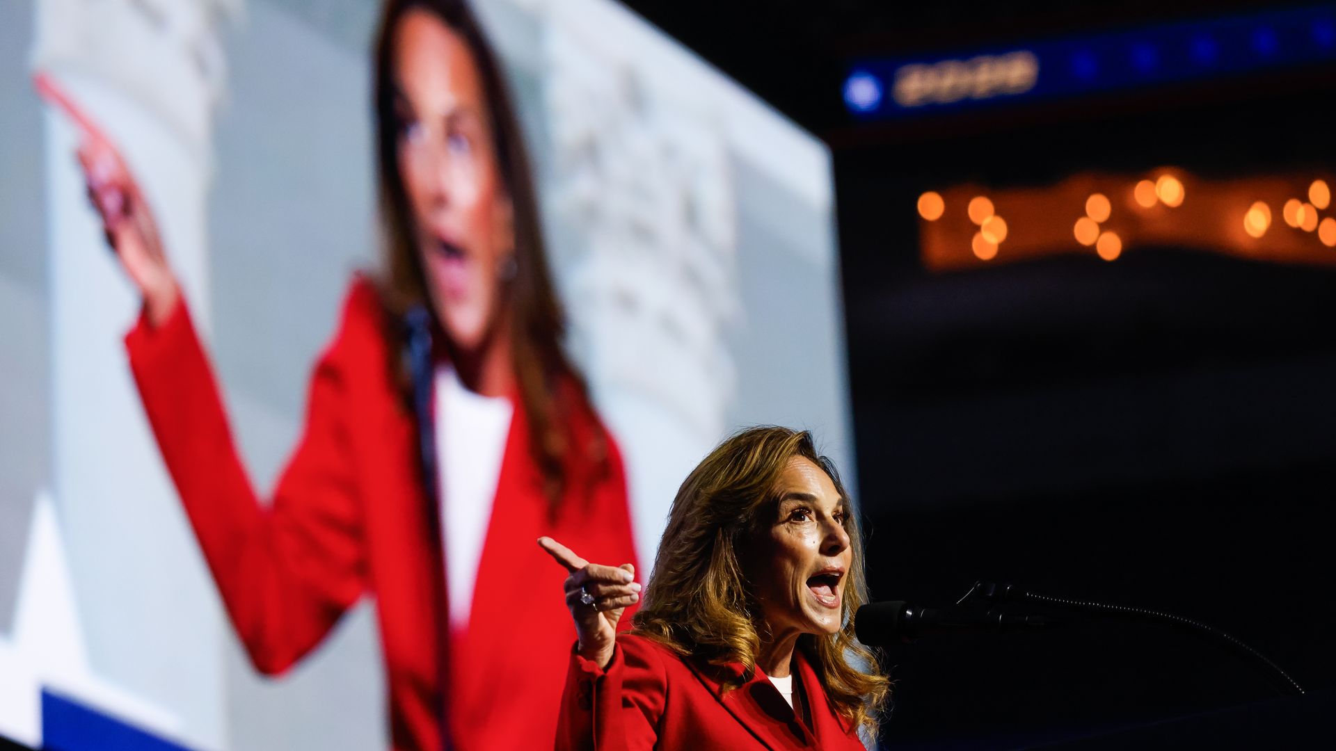 A female in a red blazer speaks at a microphone on a dark stage; a large, blurred screen behind shows another person in red, with warm orange bokeh lights in the background.