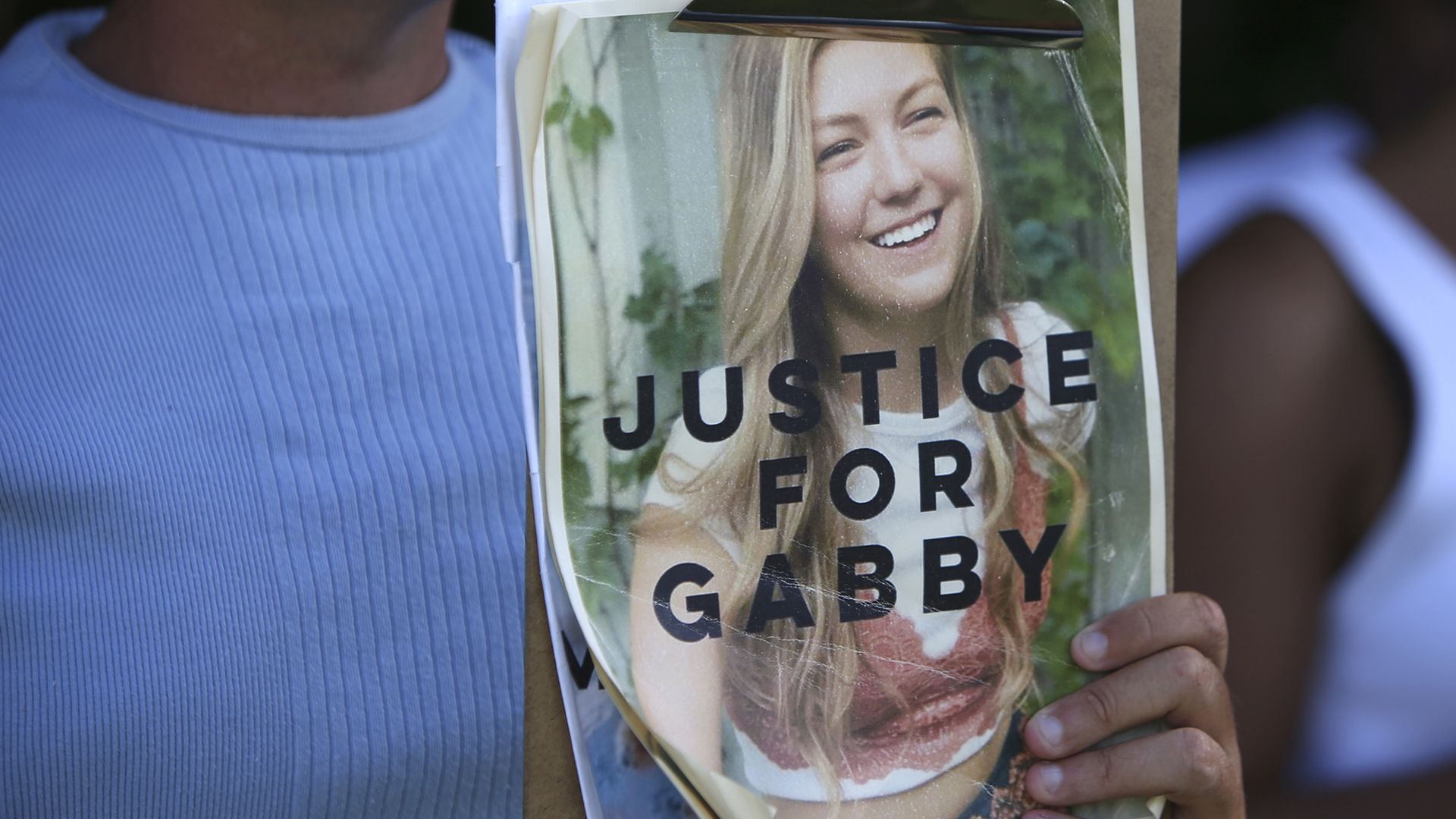 A woman holds a clipboard displaying a photo of a blonde woman with the words "Justice for Gabby" printed on the photo. 