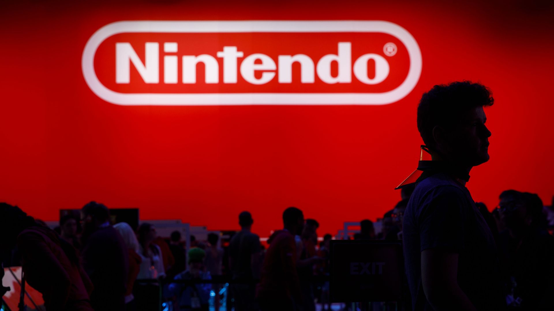 Photo showing people in a darkened conference hall in front of a large red wall bearing a white Nintendo logo