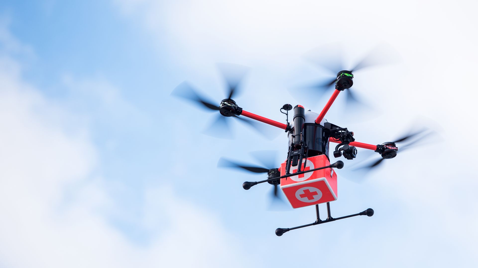 Low-angle view of a red quadcopter drone with spinning black rotor blades against a blue sky with soft clouds; a red medical/first-aid sign hangs from its frame.