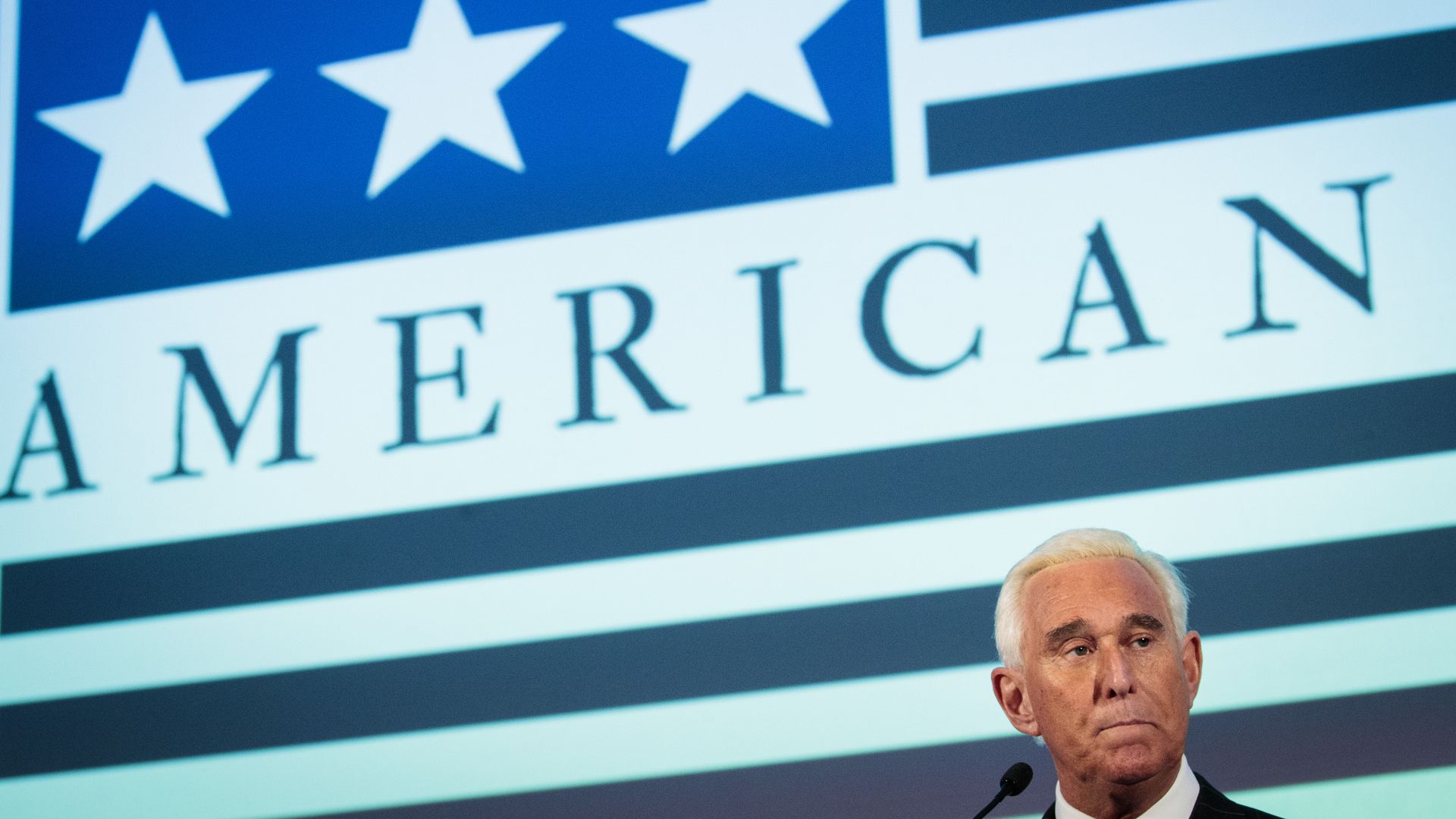 Roger Stone's head in the bottom right of the frame with a projected American flag in the back ground that says "American" on it