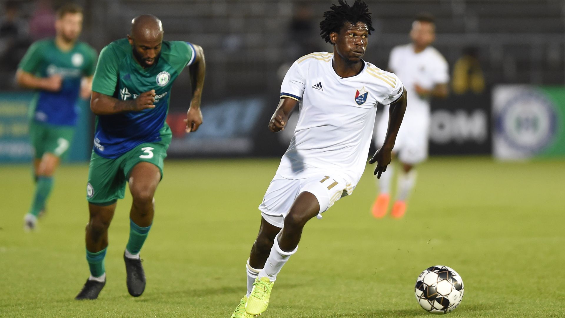 Soccer player in white jersey and neon cleats dribbles ball while an opponent in green and blue chases during a night match on a grass field with blurred spectators in the background.