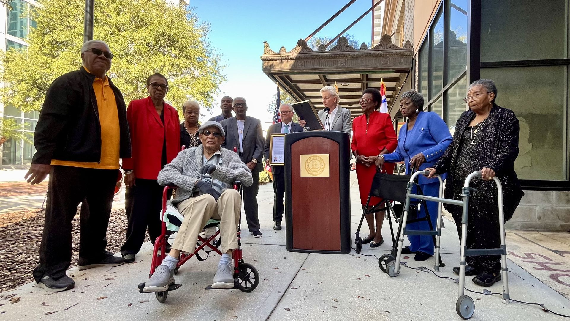 A woman in a gray suit stands at a lectern reading a framed piece of paper. She's flanked by several people, two of him are with walkers and one in a wheelchair.
