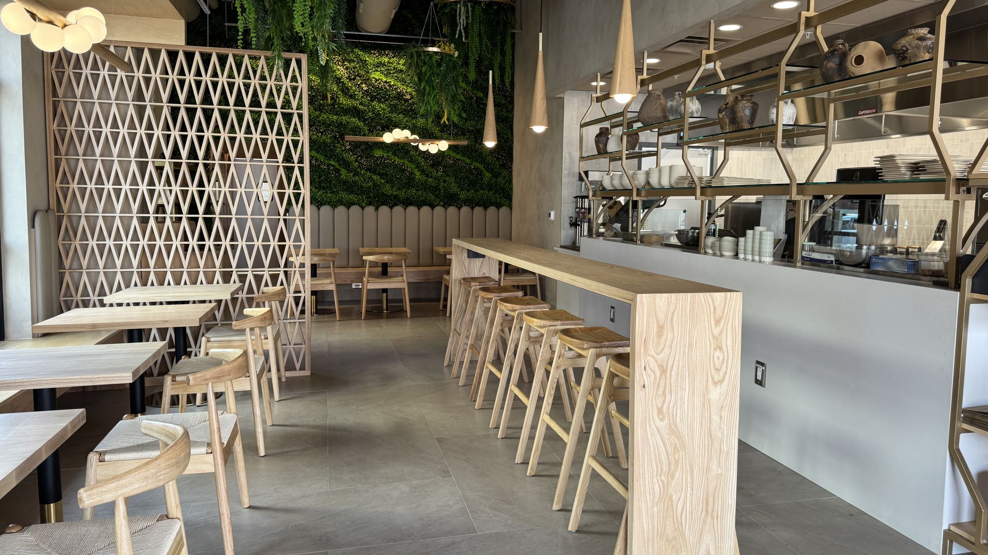 Modern cafe interior with light wood furniture, a long high table with stools, geometric wooden divider, green plant wall, hanging plants, and pendant lights above the counter and seating area.