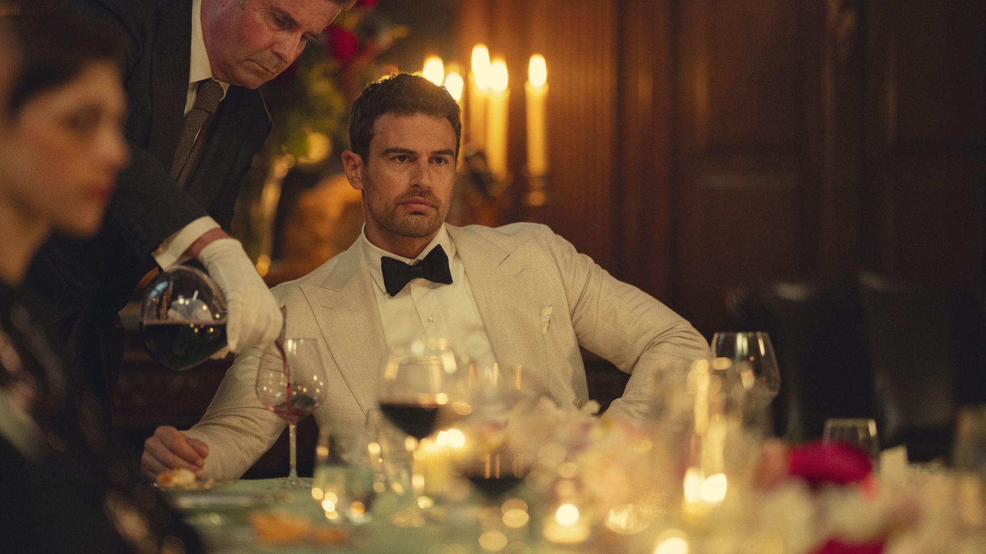 A man, Theo James, sits at a table in a tuxedo while a white-gloved hand pours him wine.