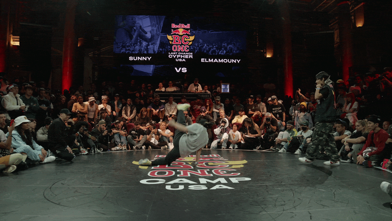 Breakdancer performs a spin in the center of a crowd-lined circle at a Red Bull BC One USA event; a large screen reads "Sunny vs Elmamouny" as spectators watch.
