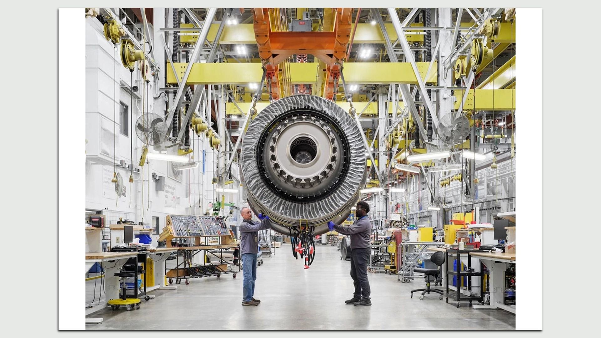Two workers in gray jackets and gloves inspecting a large aircraft engine suspended from a yellow overhead crane in a bright, spacious aerospace manufacturing facility.