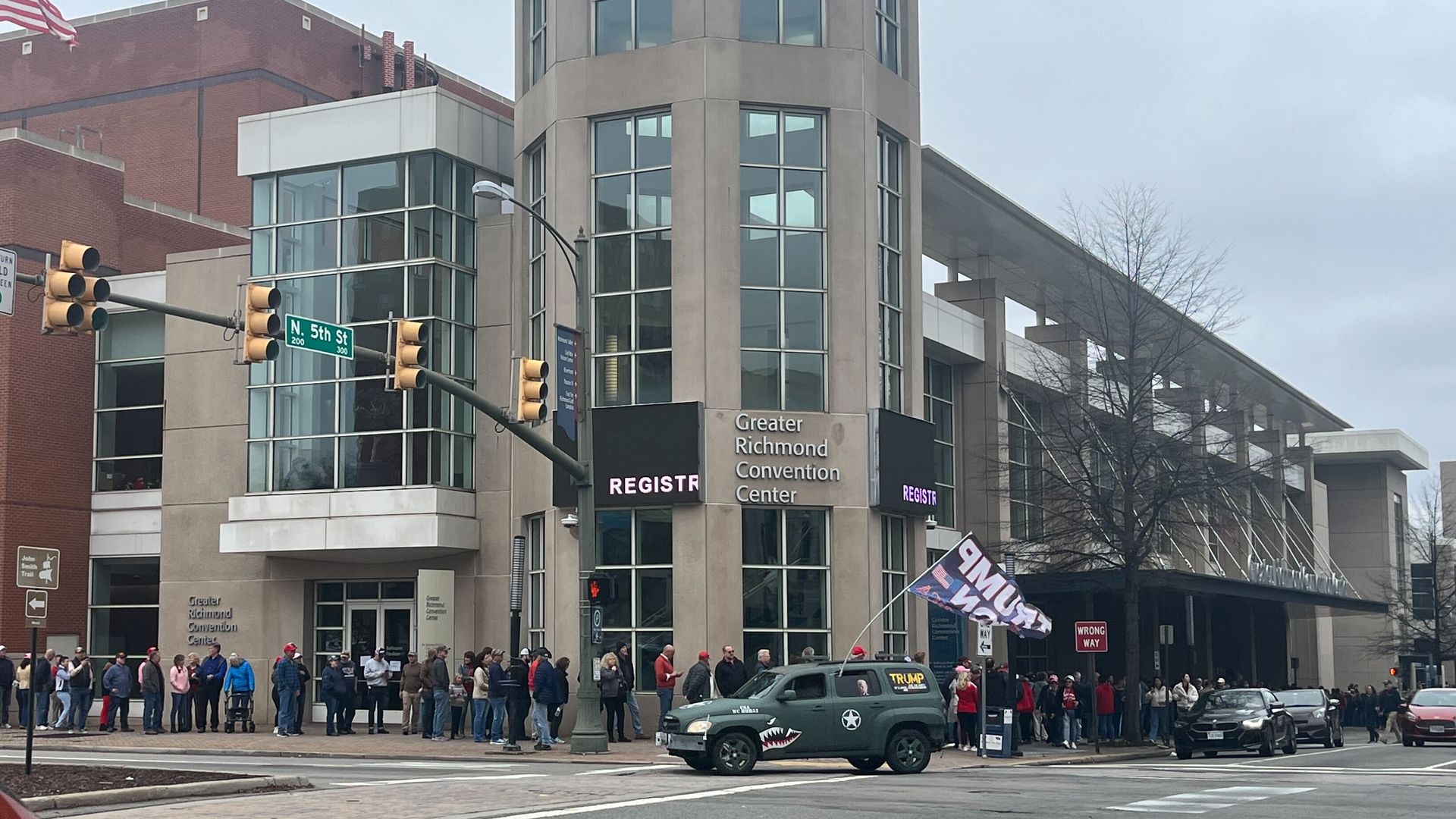 A picture of a line of people wrapping around the Greater Richmond Convention Center with a green car waving a Trump flag.
