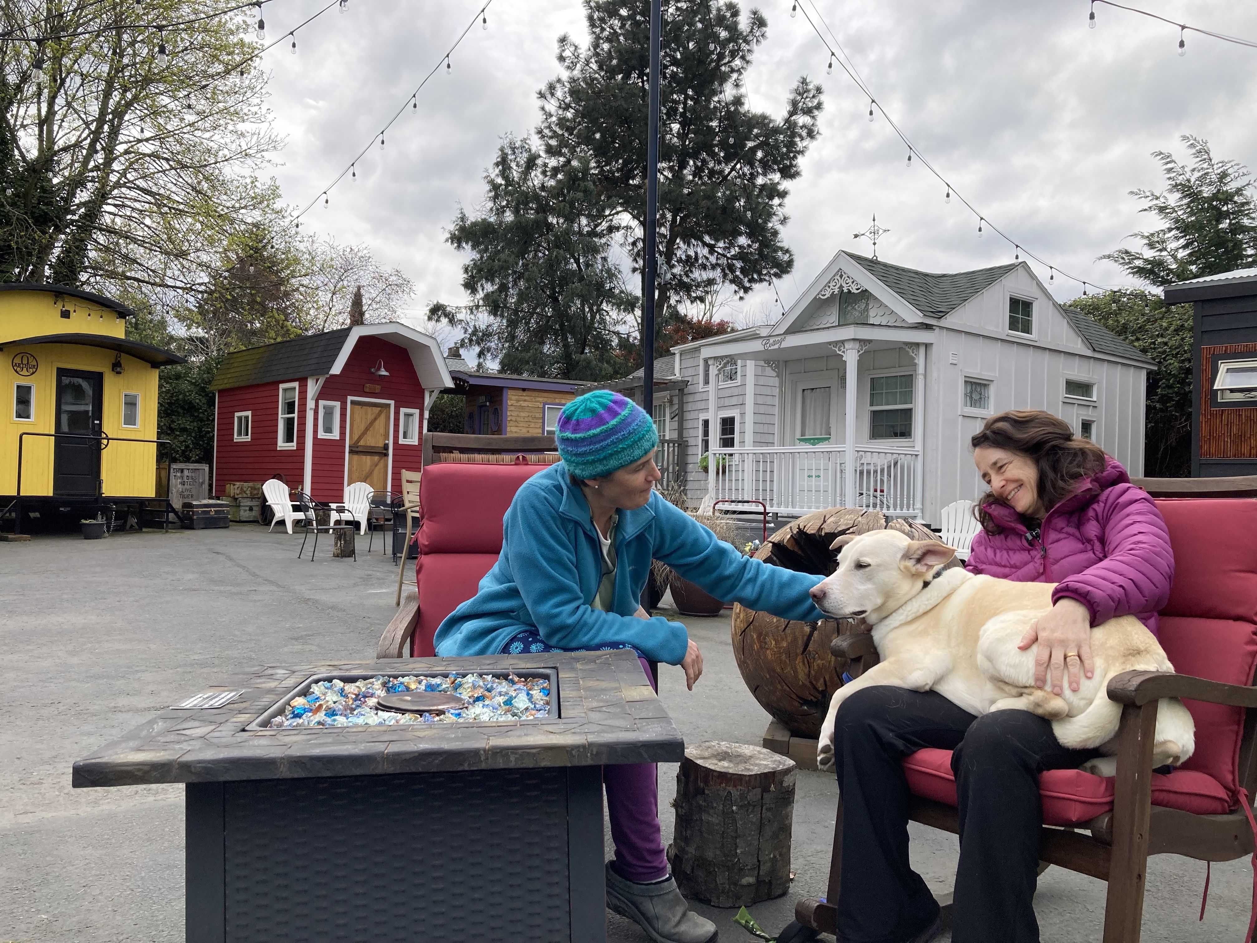 Two people sit by an outdoor fire pit, one with a dog on her lap, in an concrete yard with tiny houses are in the background.