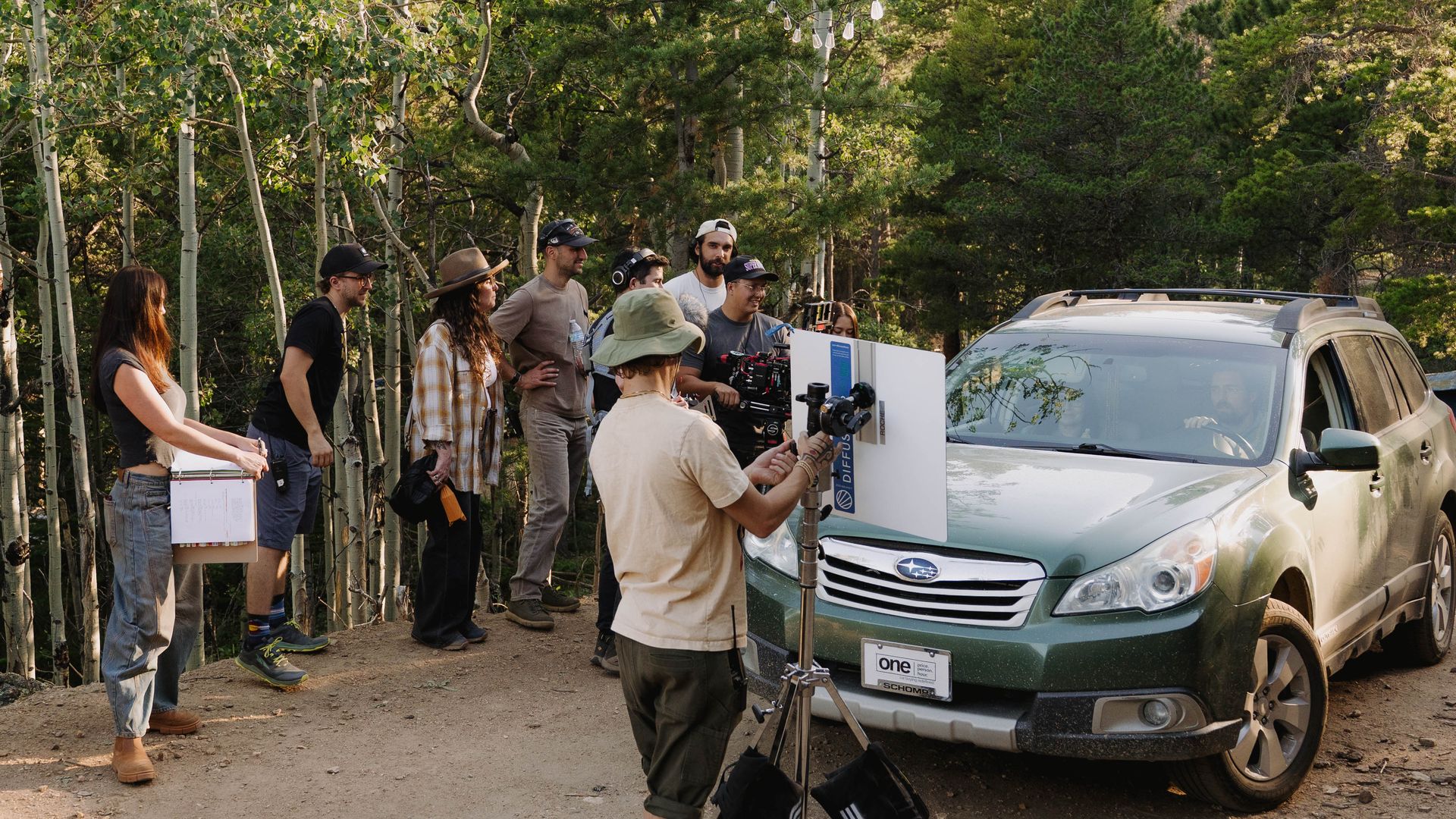 A crew of people gathers around film equipment in a forest clearing beside a silver SUV. They monitor a clapperboard and camera rig, with string lights overhead and trees in the background.