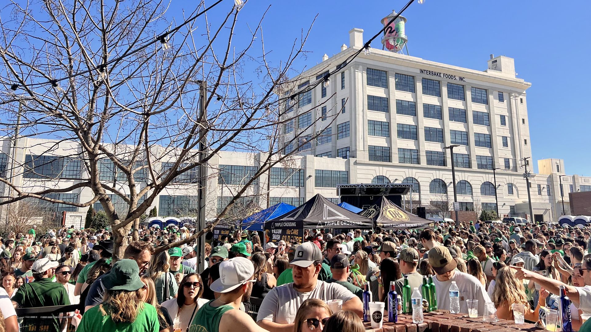 Crowd at a sunny outdoor event in front of a white building labeled "Interbake Foods, Inc." String lights cross the blue sky; a leafless tree sits to the left. Tents and a brick bar fill the foreground