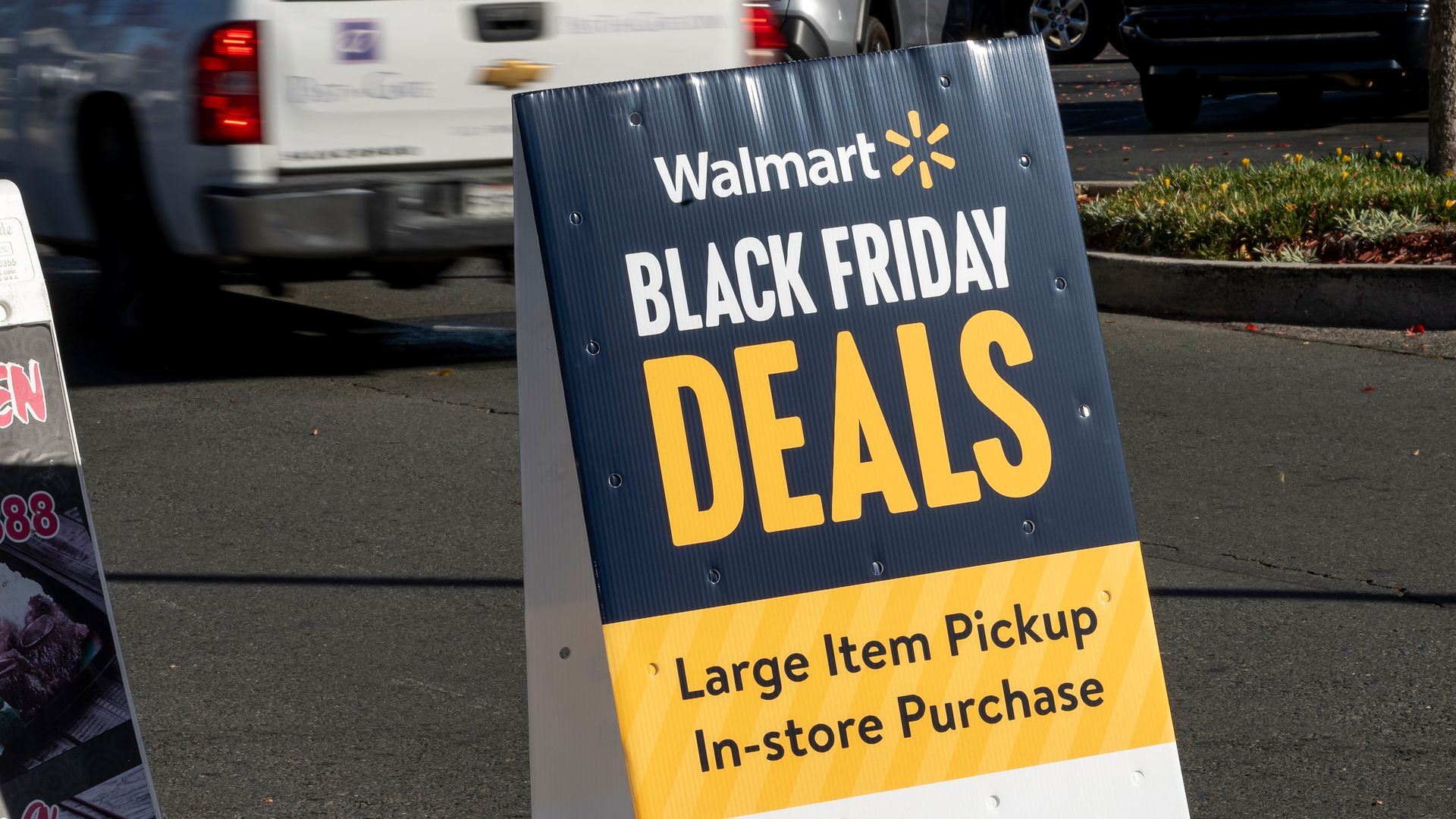A Walmart sign on a parking lot advertising Black Friday deals with a note about large item pickup for in-store purchases, surrounded by cars and pavement.