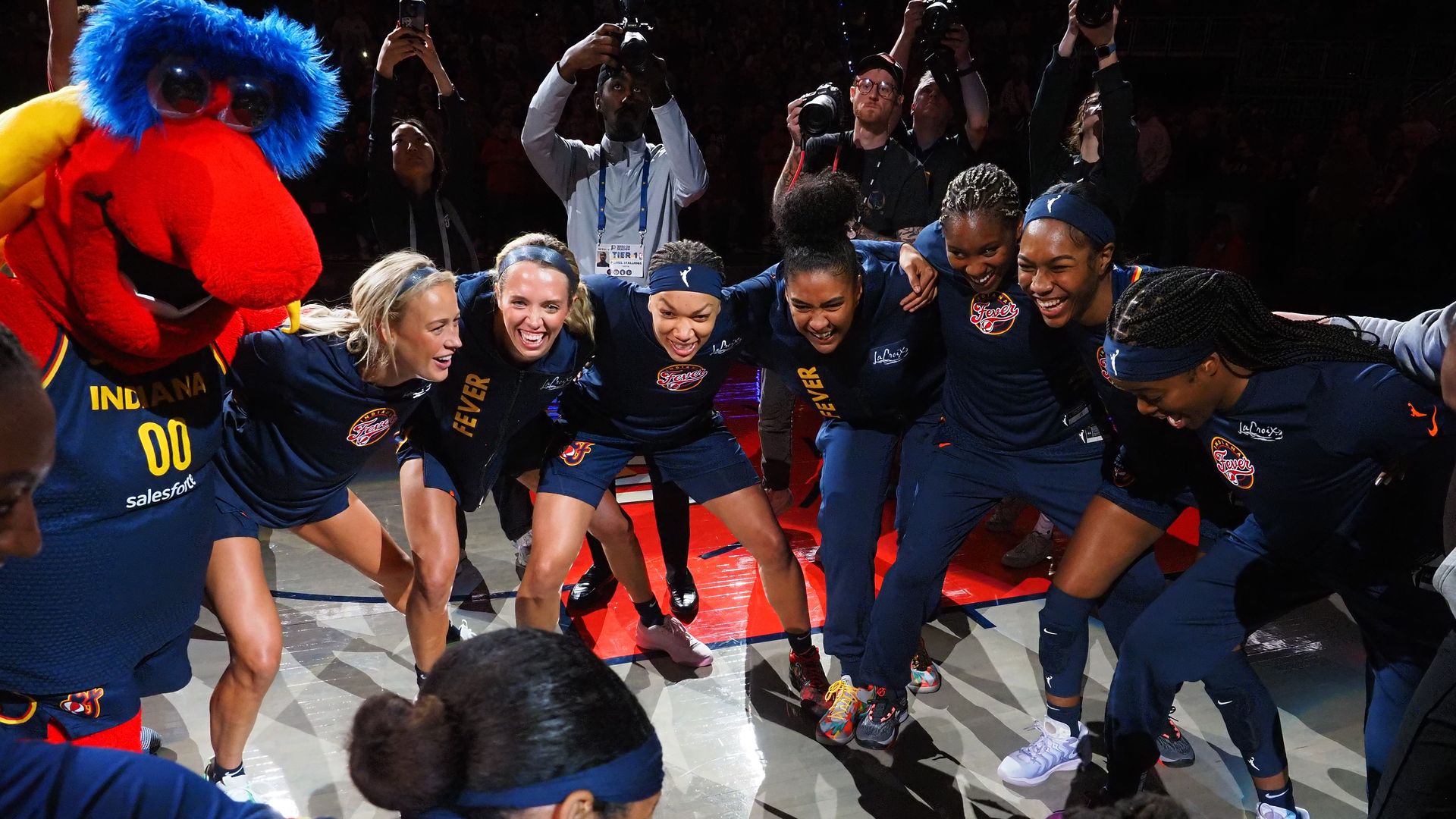 Indiana Fever women's basketball players and mascot in a huddle on court, wearing navy and yellow uniforms, with photographers capturing the moment in the background.