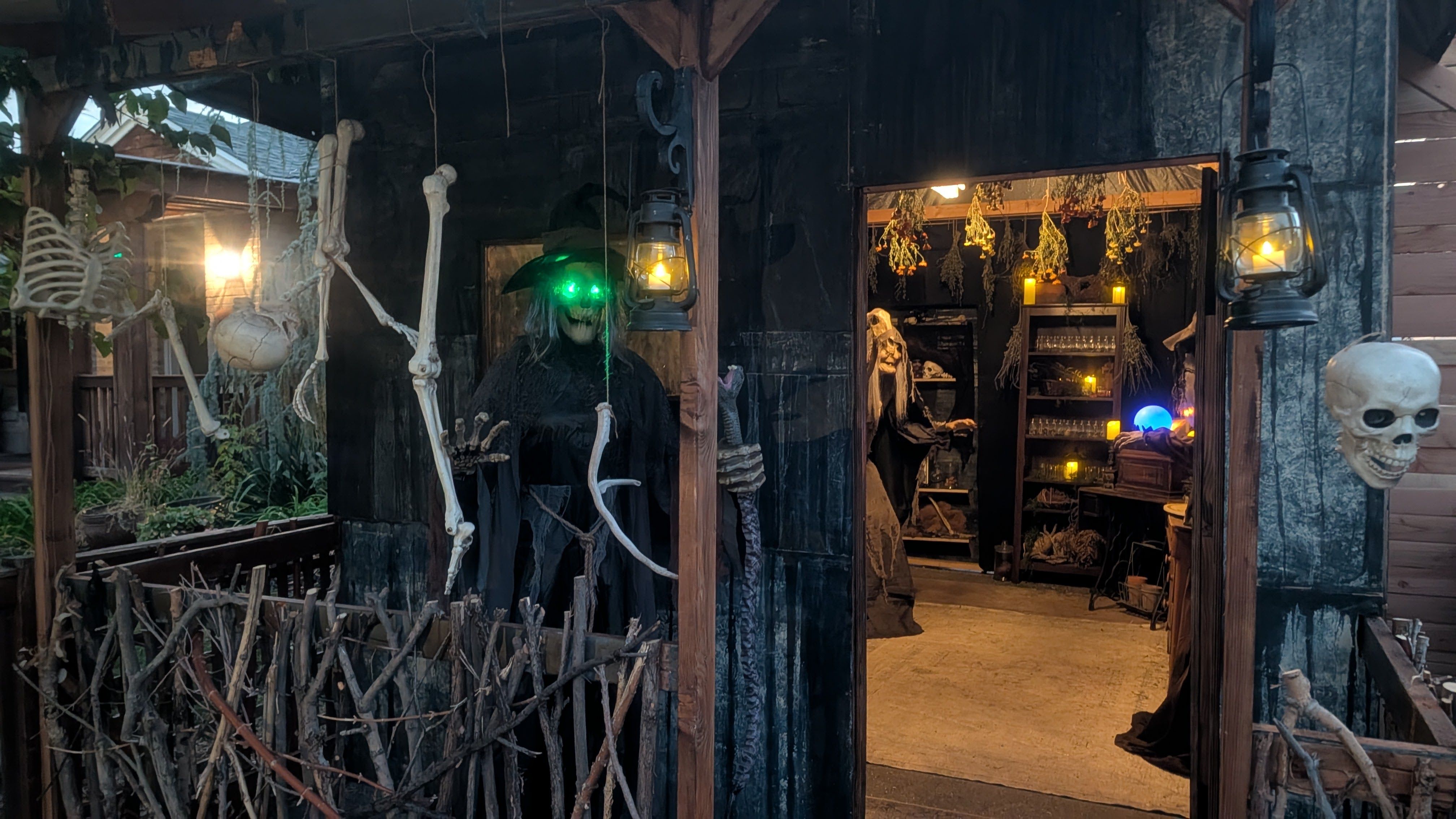 Dark wooden Halloween shop facade with hanging skeletons and bones. Warm lanterns glow inside; a figure with green glowing eyes stands by the doorway, while skulls adorn the posts.