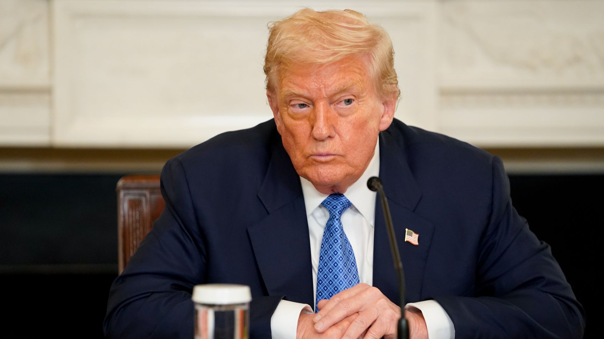 President Trump wearing a dark blue suit, white shirt, and blue patterned tie sits at a table with a microphone, a glass of water, and documents, against a neutral background.