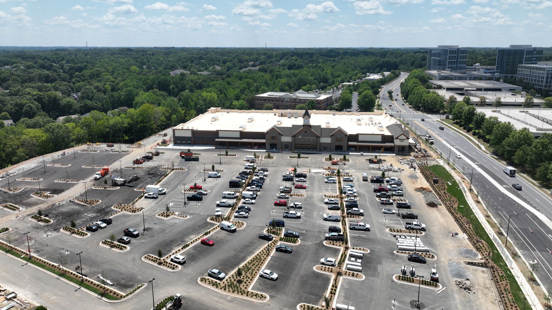 An aerial daytime view of a Wegmans store with a large parking lot, dense green trees, modern glass buildings to the right, and a busy road, under a blue sky with scattered white clouds.