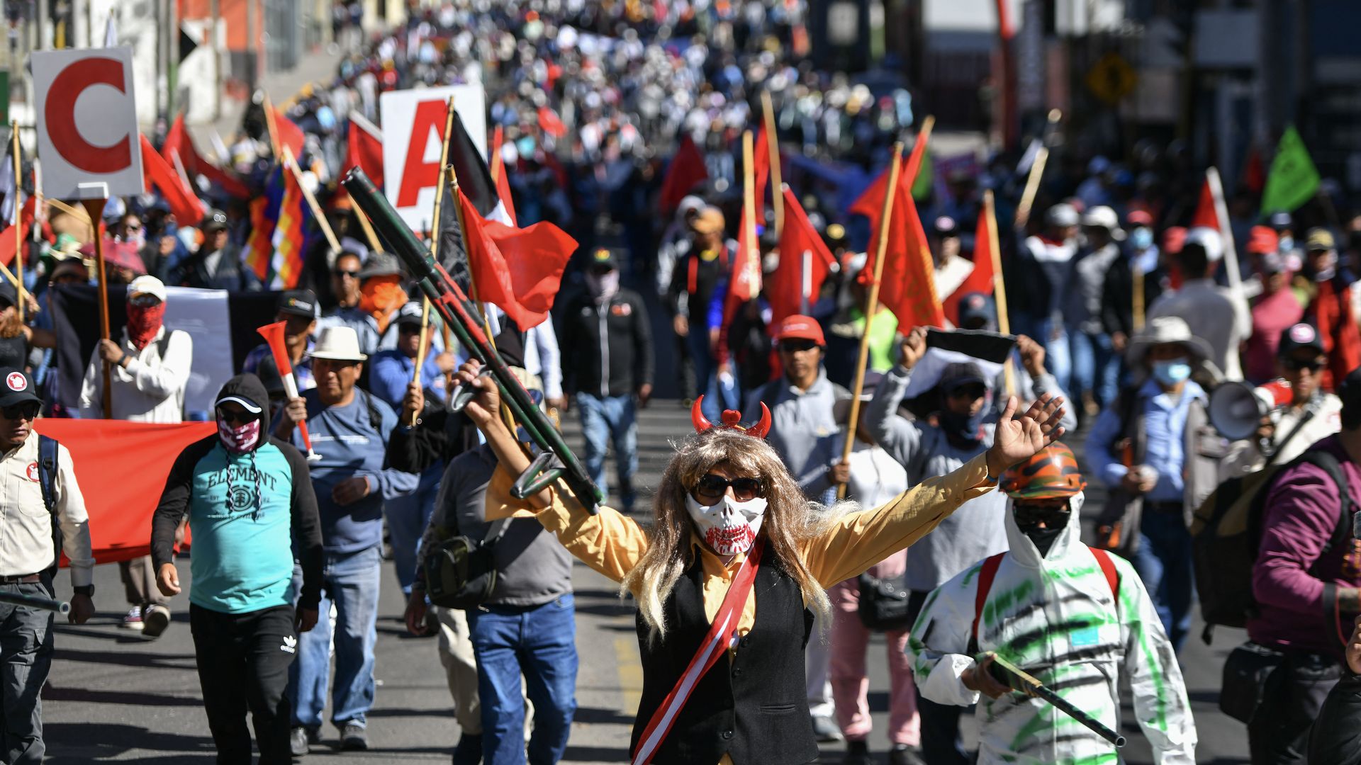 Dozens of proteters walk in the sun in Peru. One person at the front is wearing a blond wig, mask and holding up an object