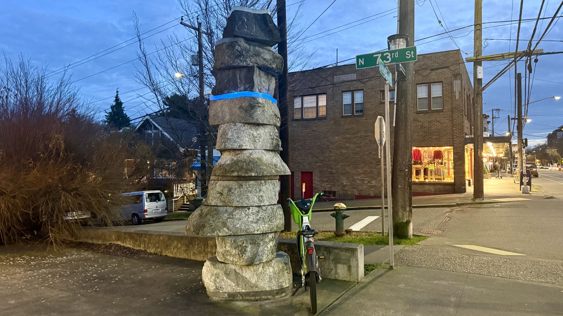 Evening street scene at N 73rd St with a stacked rock sculpture tied with a blue band next to a green bike, brick building and lit storefront in the background under a blue sky.