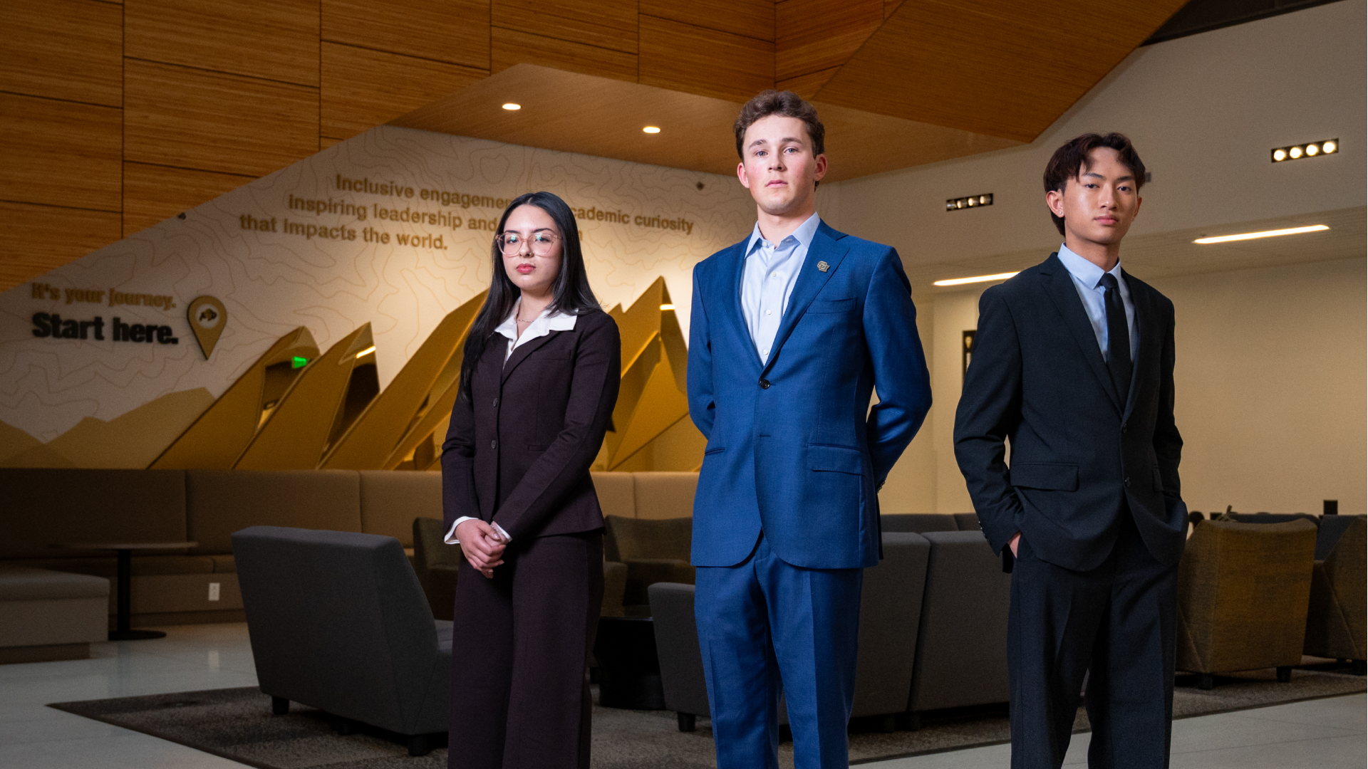 Three young professionals in formal suits stand in a modern, warmly lit lobby with wooden paneling. A wall behind them reads "Start here" with inspirational text.