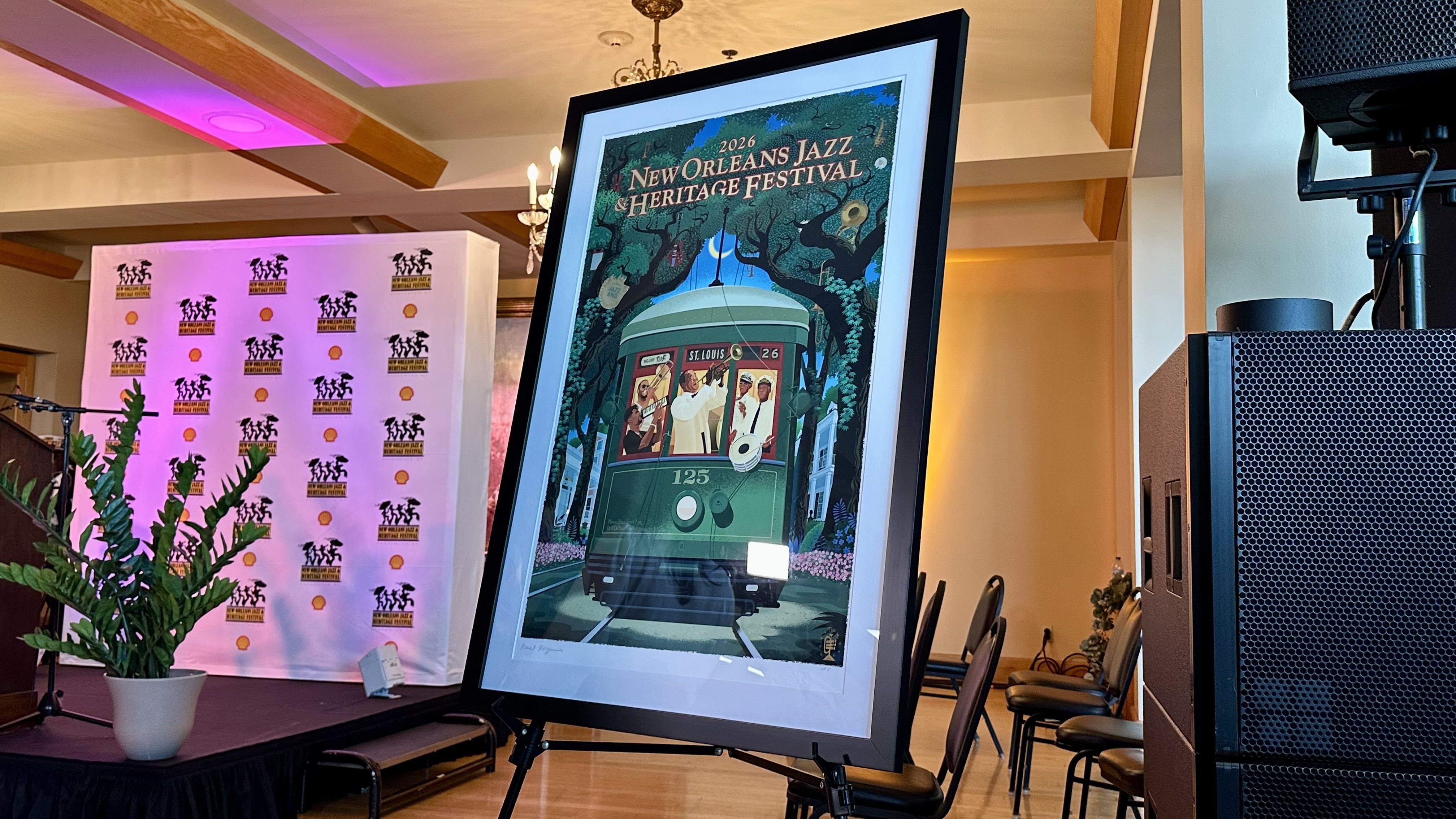 Framed poster on a stand shows "2026 New Orleans Jazz Heritage Festival" with a green streetcar under trees; pink stage lights, a logos backdrop, potted plant, and audio gear.