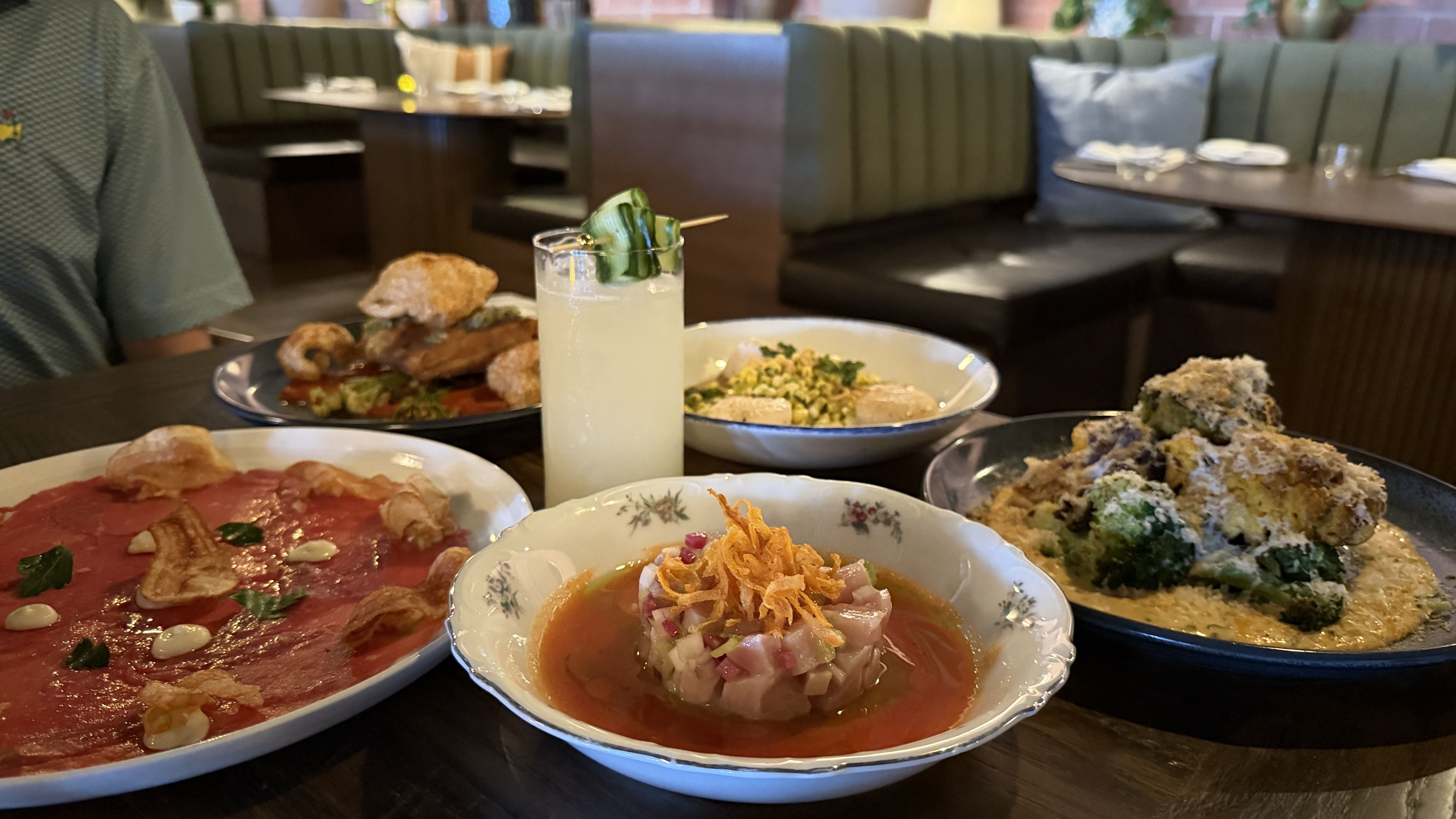 Table in a cozy restaurant with many dishes: a pale yellow cocktail with cucumber, a red meat carpaccio plate, a bowl of mixed greens, and a crispy fried platter, on a dark wooden table.