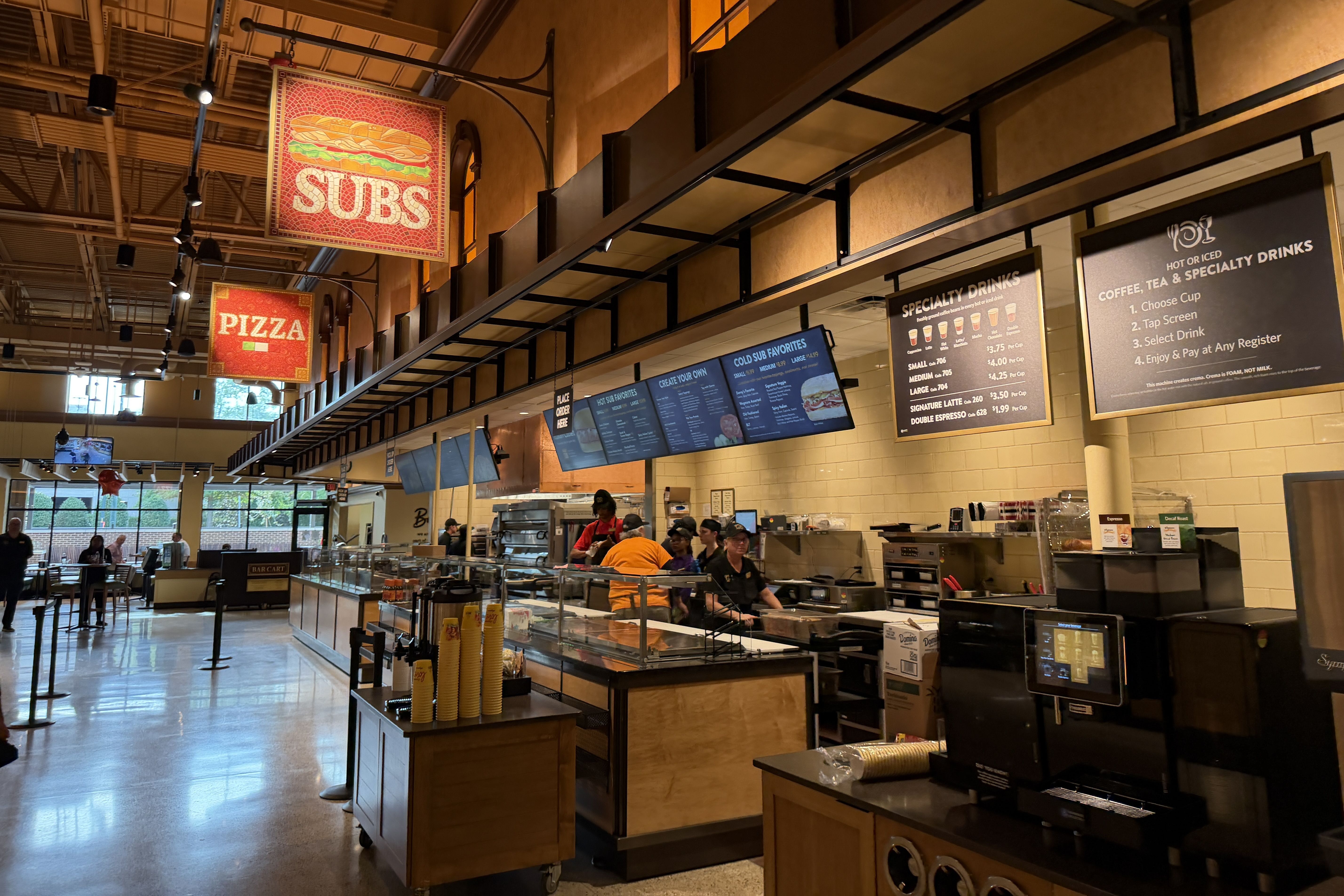 Inside a busy food hall with orange lighting, large red signs say "SUBS" and "PIZZA" above a long service counter. Menu boards and staff visible; diners sit at tables in the background.